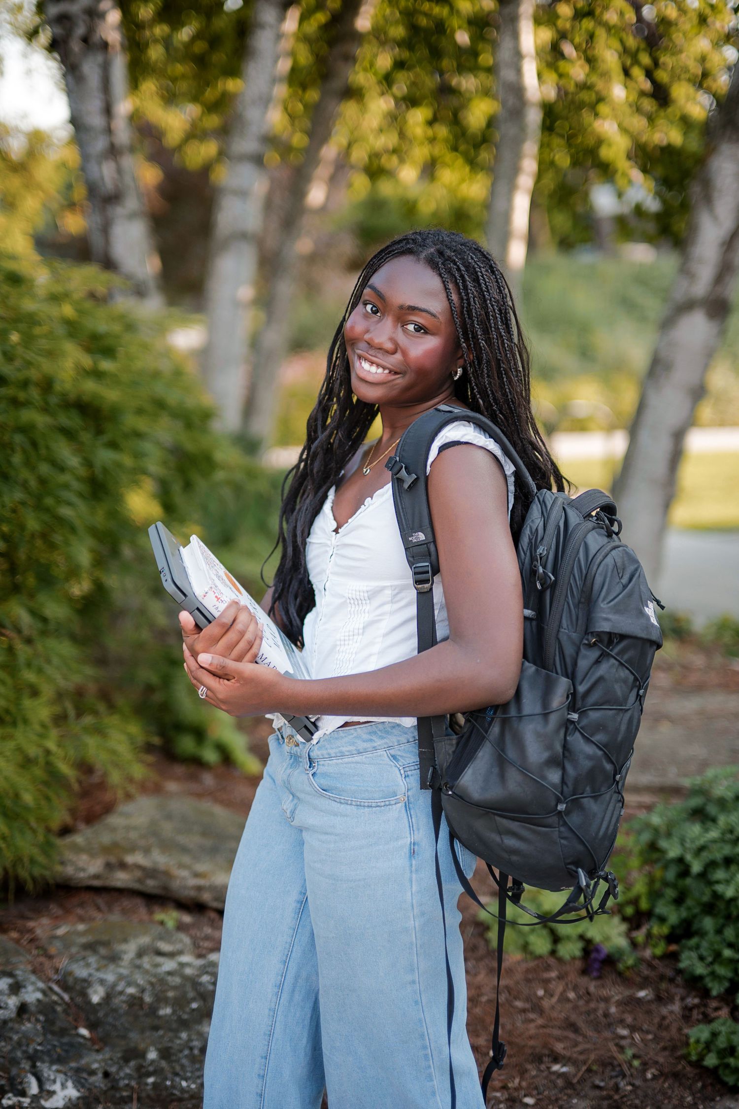 Smiling young woman with long braids carrying a black backpack and books while standing outdoors among trees.