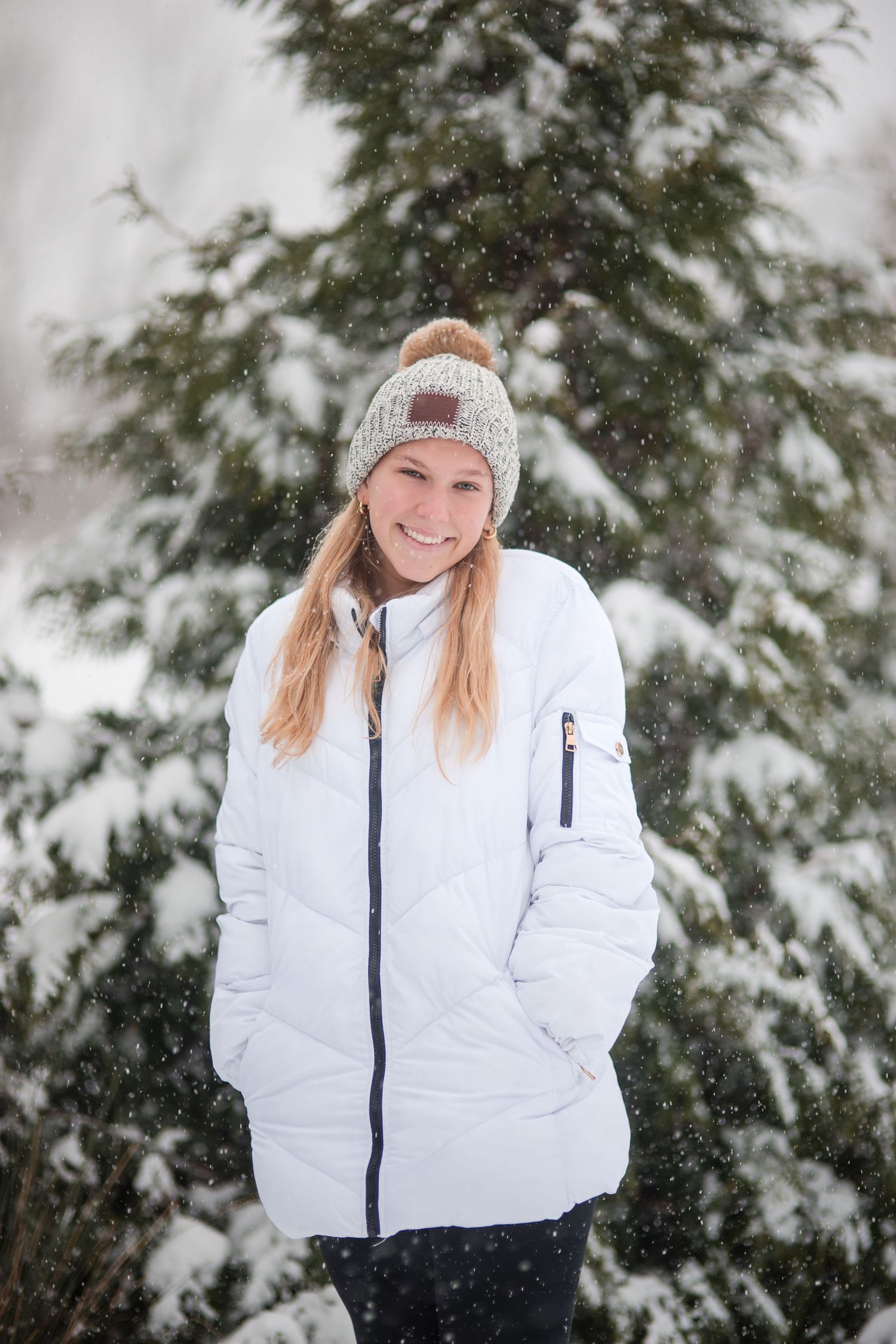 Smiling woman in a white puffer jacket and gray knit pom hat stands in front of snow-covered evergreen trees during light snowfall.