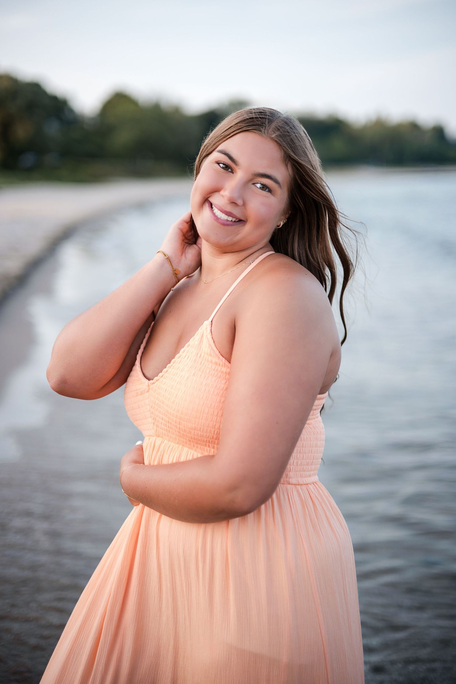 Smiling young woman in a peach dress posing by a sandy beach shoreline at golden hour.