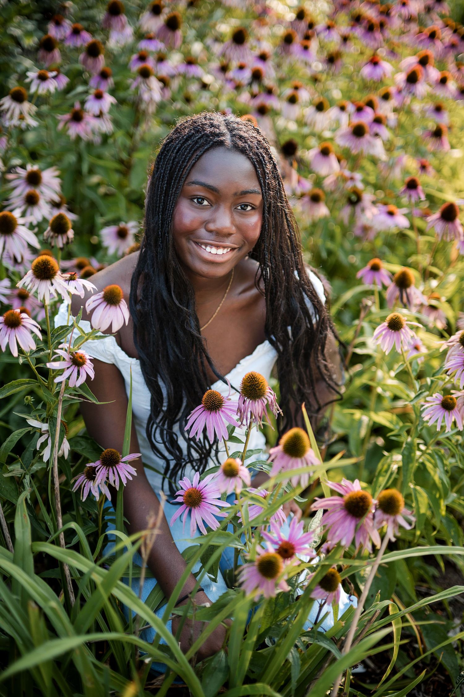 Smiling young woman with long braids in a white dress sits among blooming purple coneflowers in a sunlit garden.