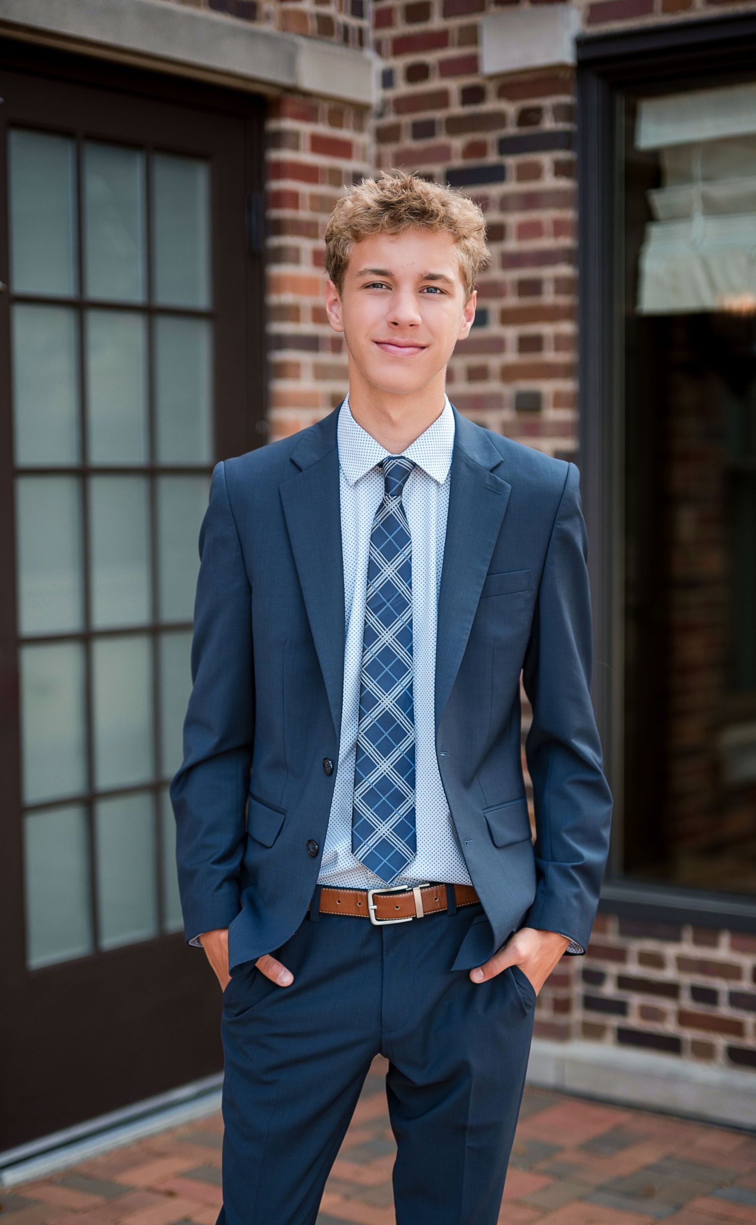 Smiling teenage boy in a navy blazer, plaid tie and brown leather belt standing with hands in pockets outside a brick building.