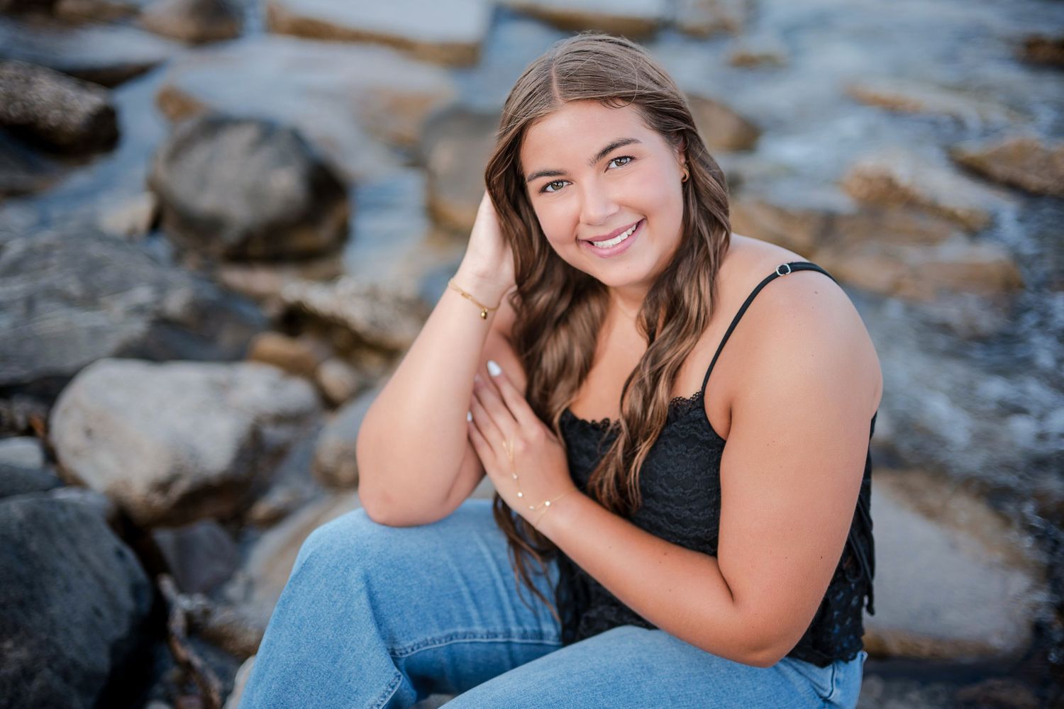 Young woman with long brown hair smiling on rocky beach, wearing black top and jeans in a senior portrait.