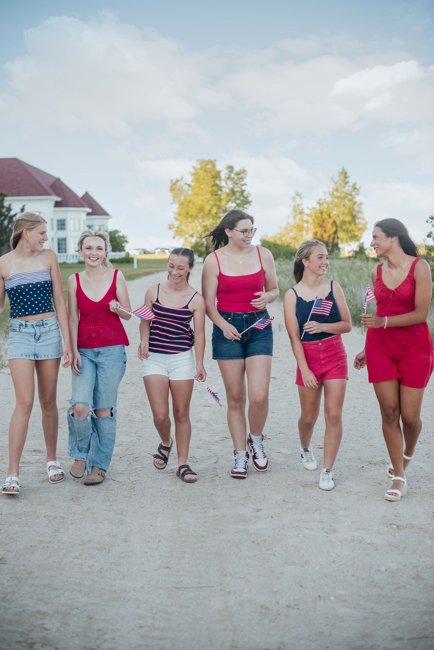 Group of six young women in red, white, and blue outfits walking together on a sandy path while holding small American flags.