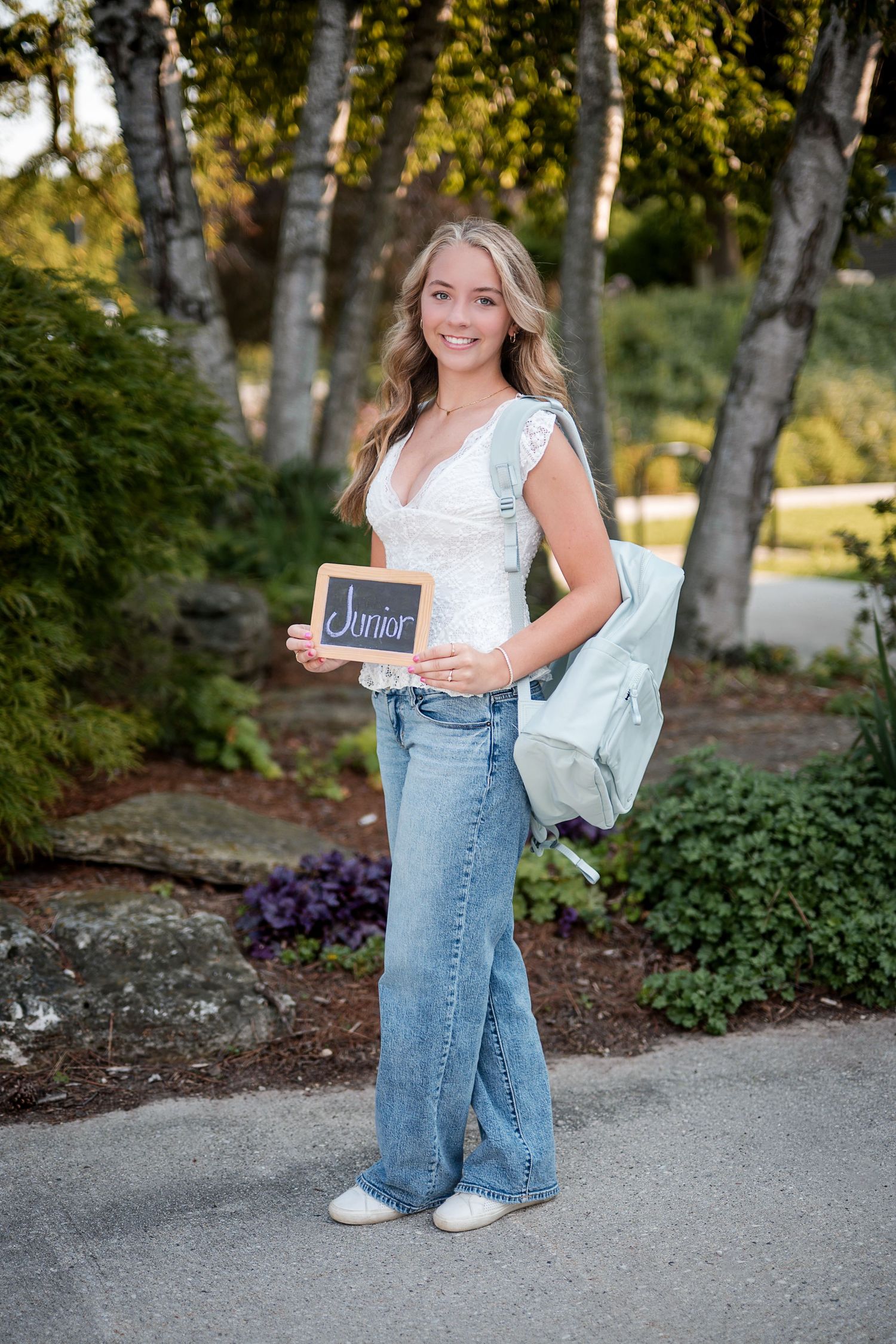 Smiling teen girl in a white top and jeans holds a small chalkboard reading Junior and wears a mint backpack outdoors.