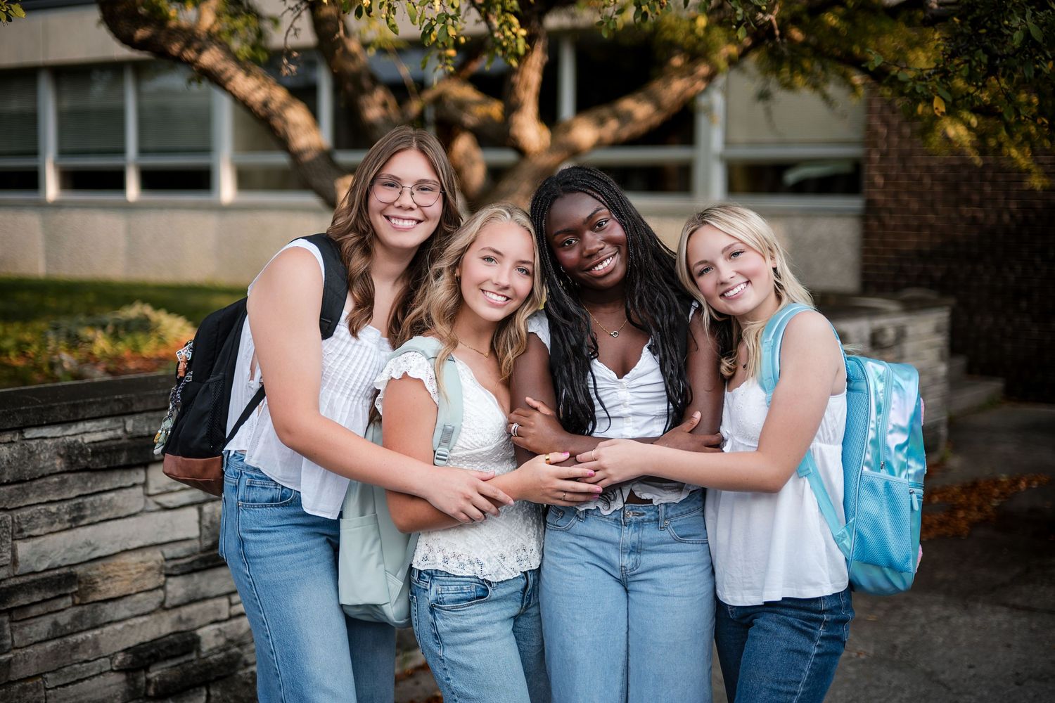 Four smiling teenage girls with backpacks posing arm in arm outdoors near a stone wall and trees on a school campus.