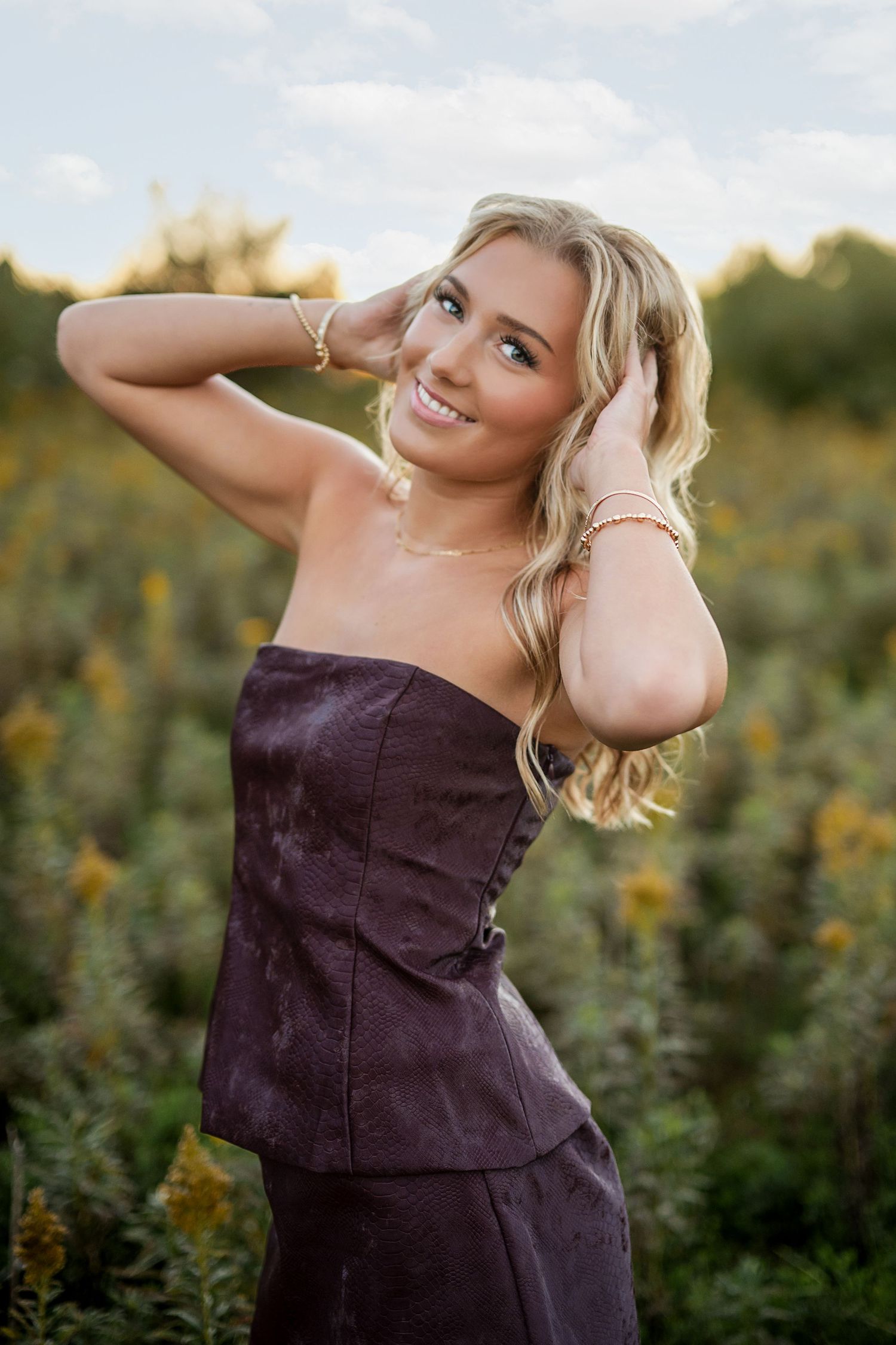 Smiling blonde woman in a purple strapless top poses with her hands in her hair in a field of yellow wildflowers.