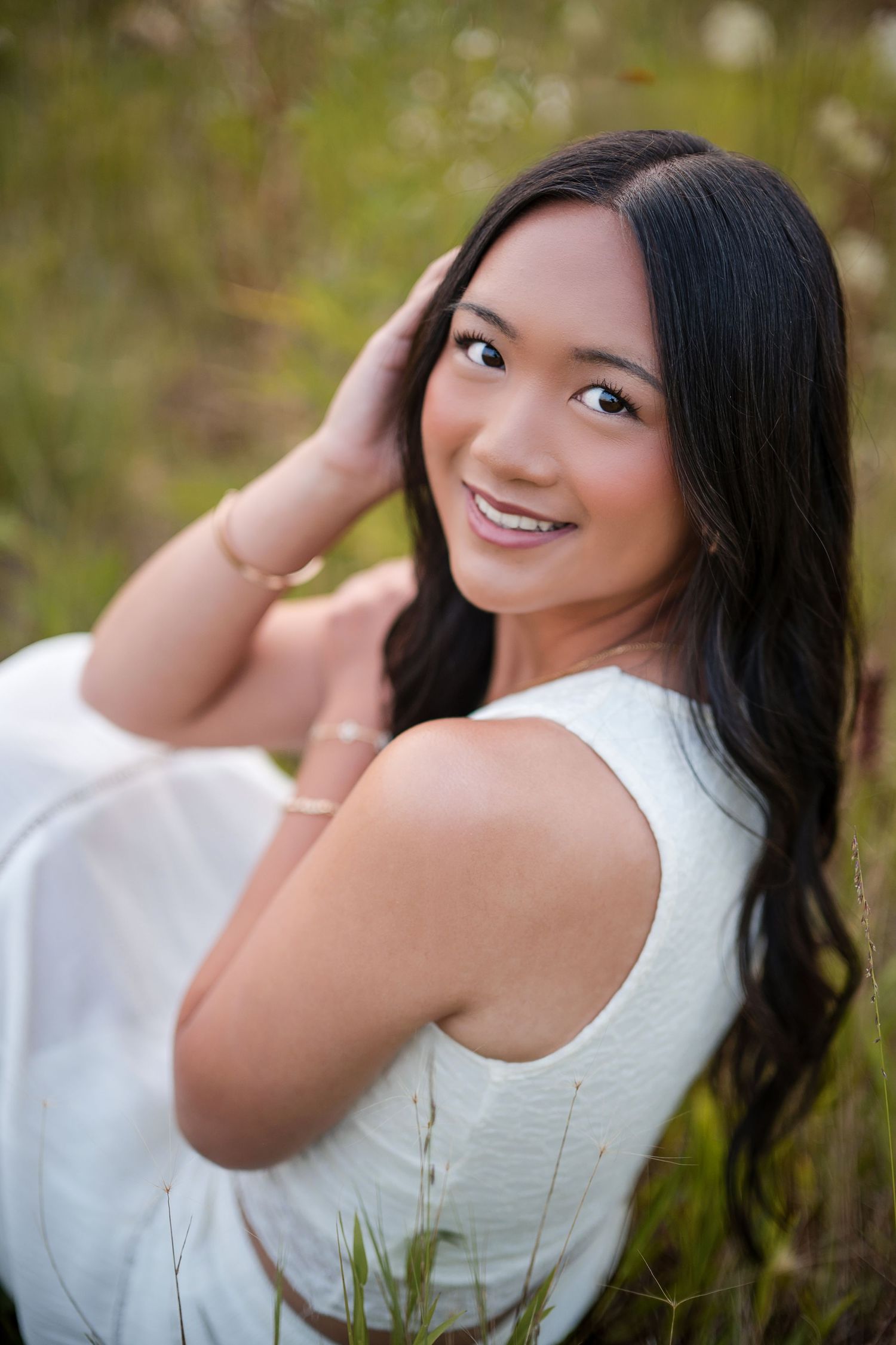 Smiling young woman with long dark hair in white dress sitting outdoors in a natural green field setting.
