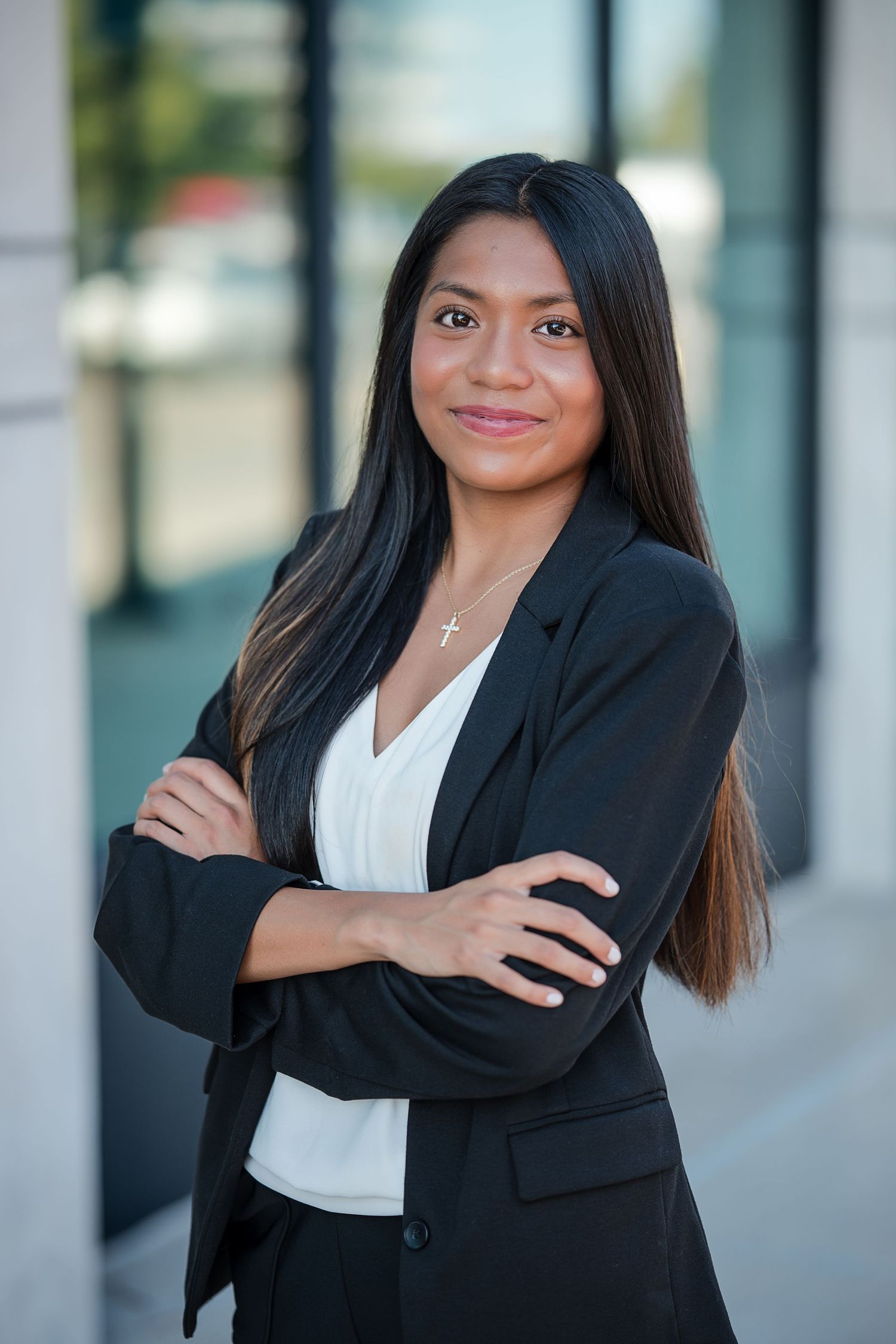 Young businesswoman in black blazer stands confidently with arms crossed, smiling outdoors near modern building.