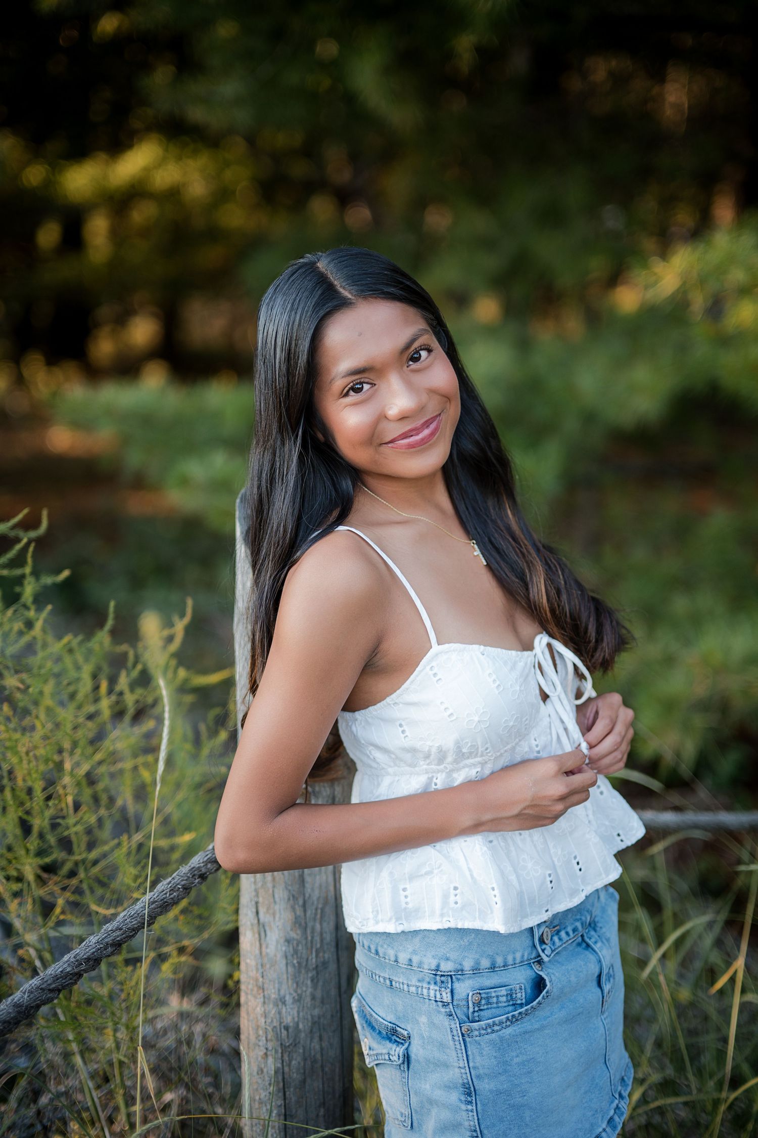 Young woman in white eyelet top and jeans smiles while leaning against a wooden fence post outdoors.
