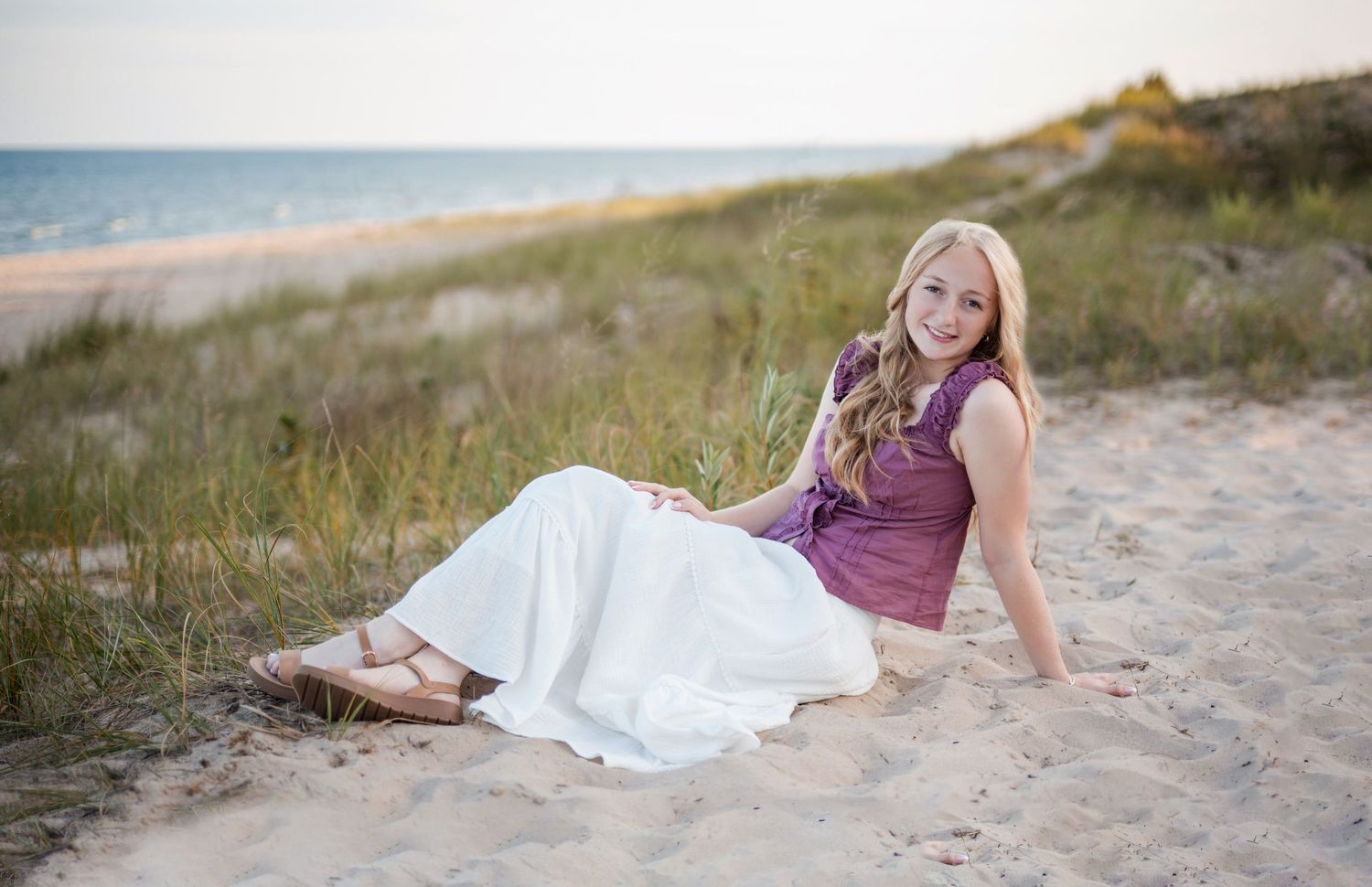 Smiling Senior girl wearing a purple top and white skirt sitting on sandy beach dunes with at Terry Andrae State Park along Lake Michigan