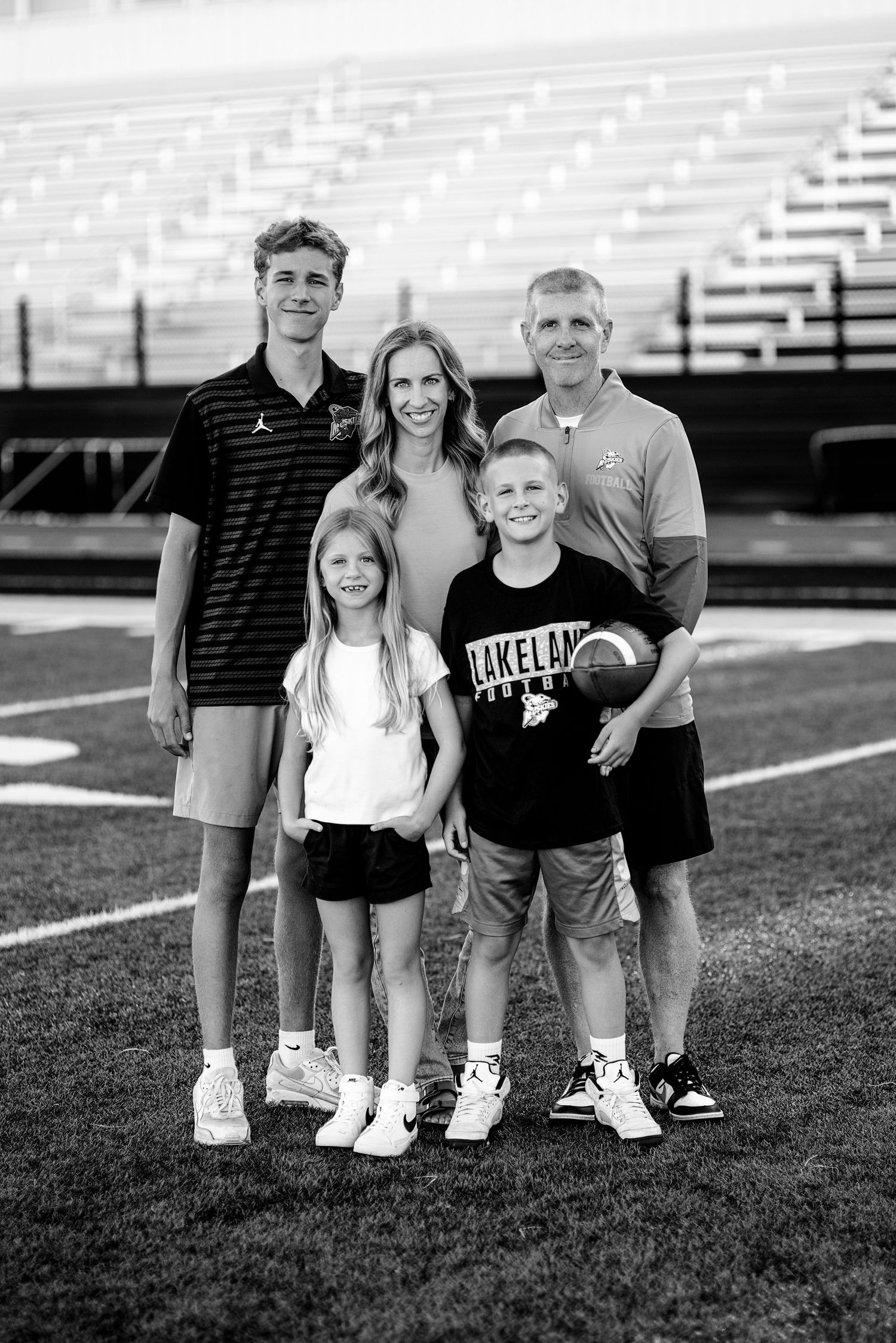 Black-and-white family portrait of two parents and three children, one holding a football, standing on a stadium field.