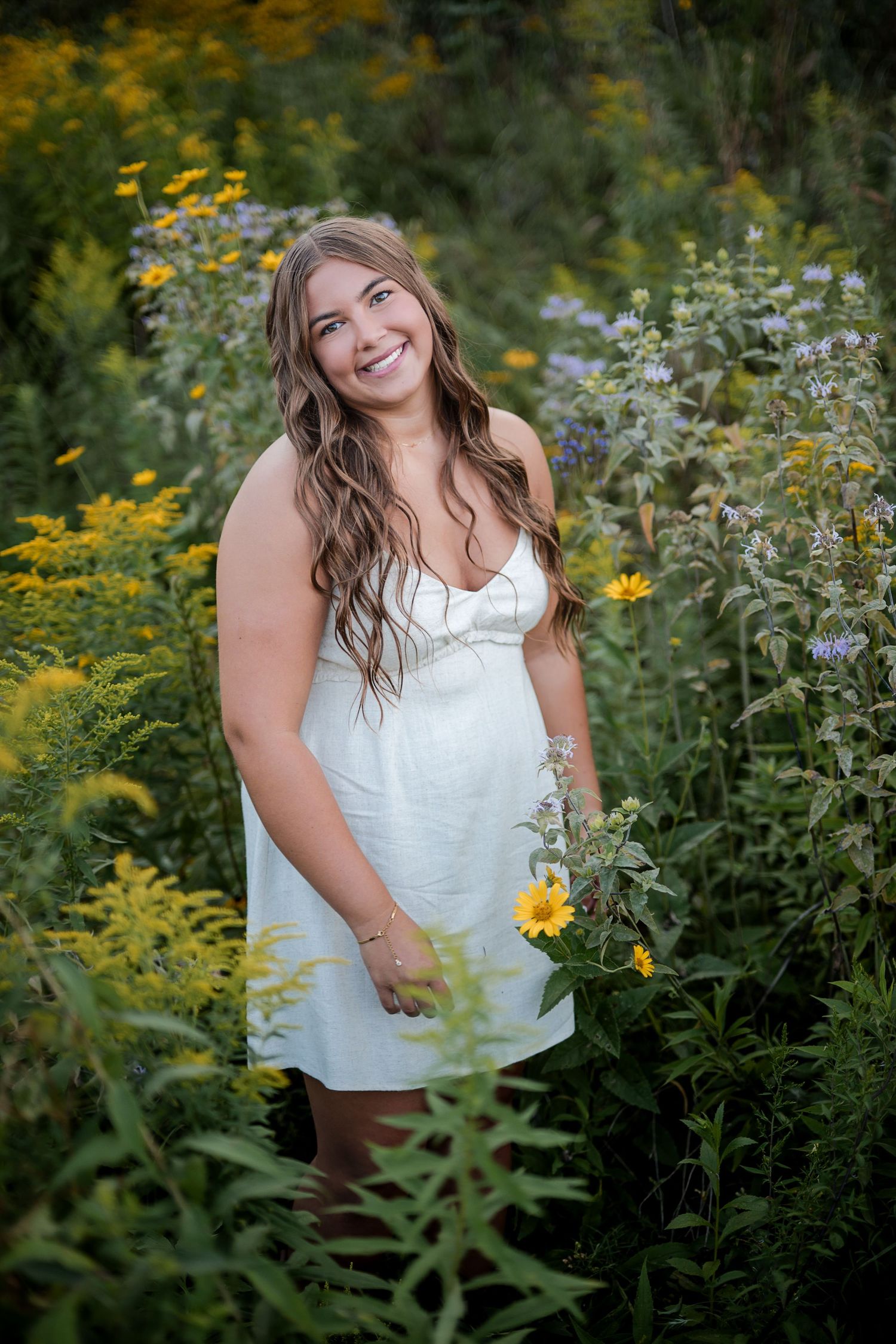 Smiling young woman in white dress standing among yellow and purple wildflowers in a lush garden setting.