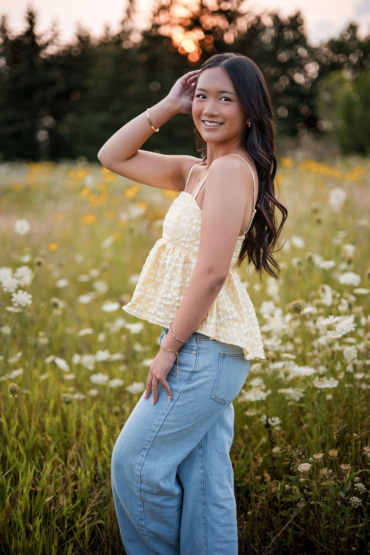 Young woman in yellow top and jeans smiling in a wildflower field at golden hour sunset.