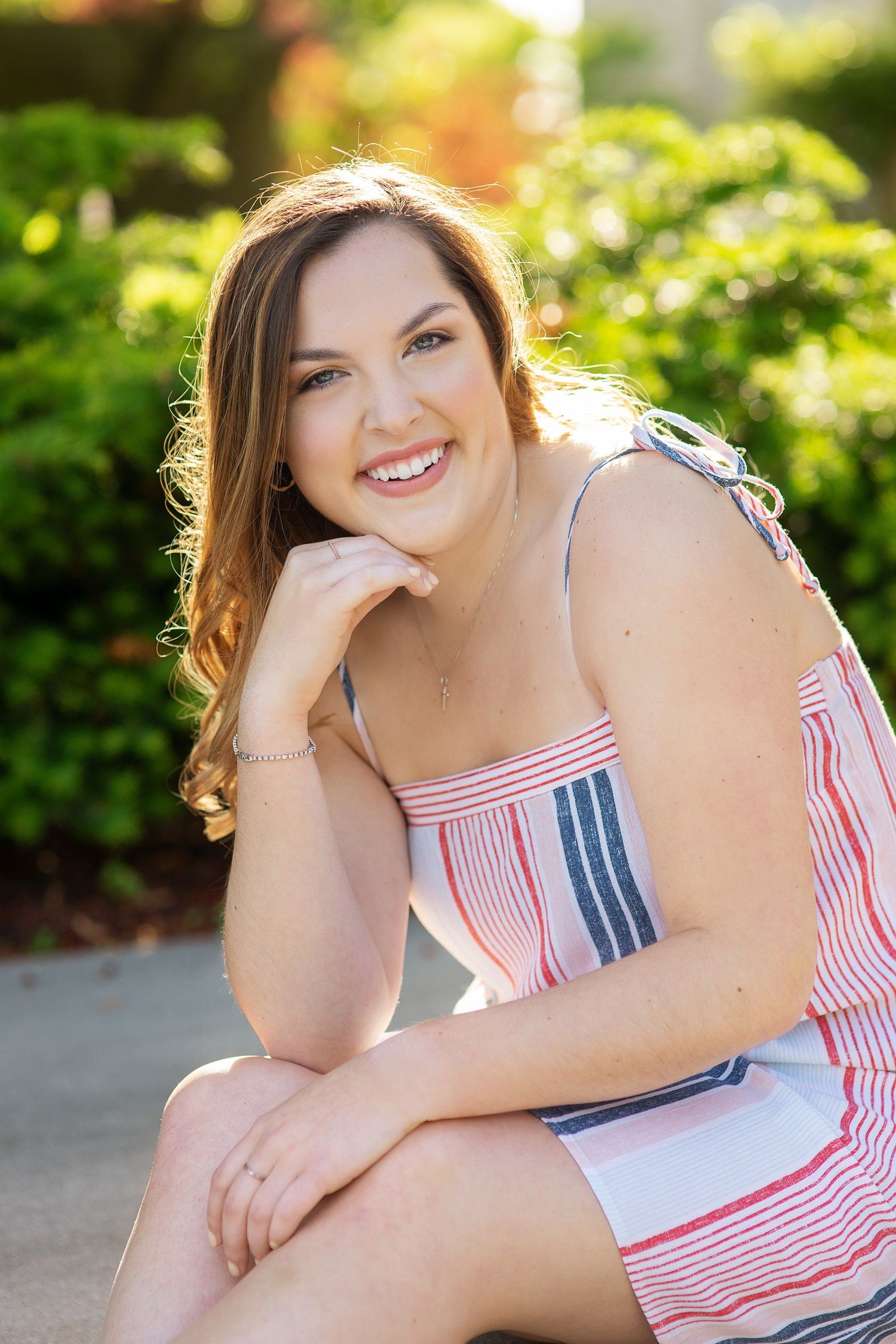 Smiling young woman with long brown hair in a striped sundress rests her chin on her hand while sitting outdoors near green shrubs.