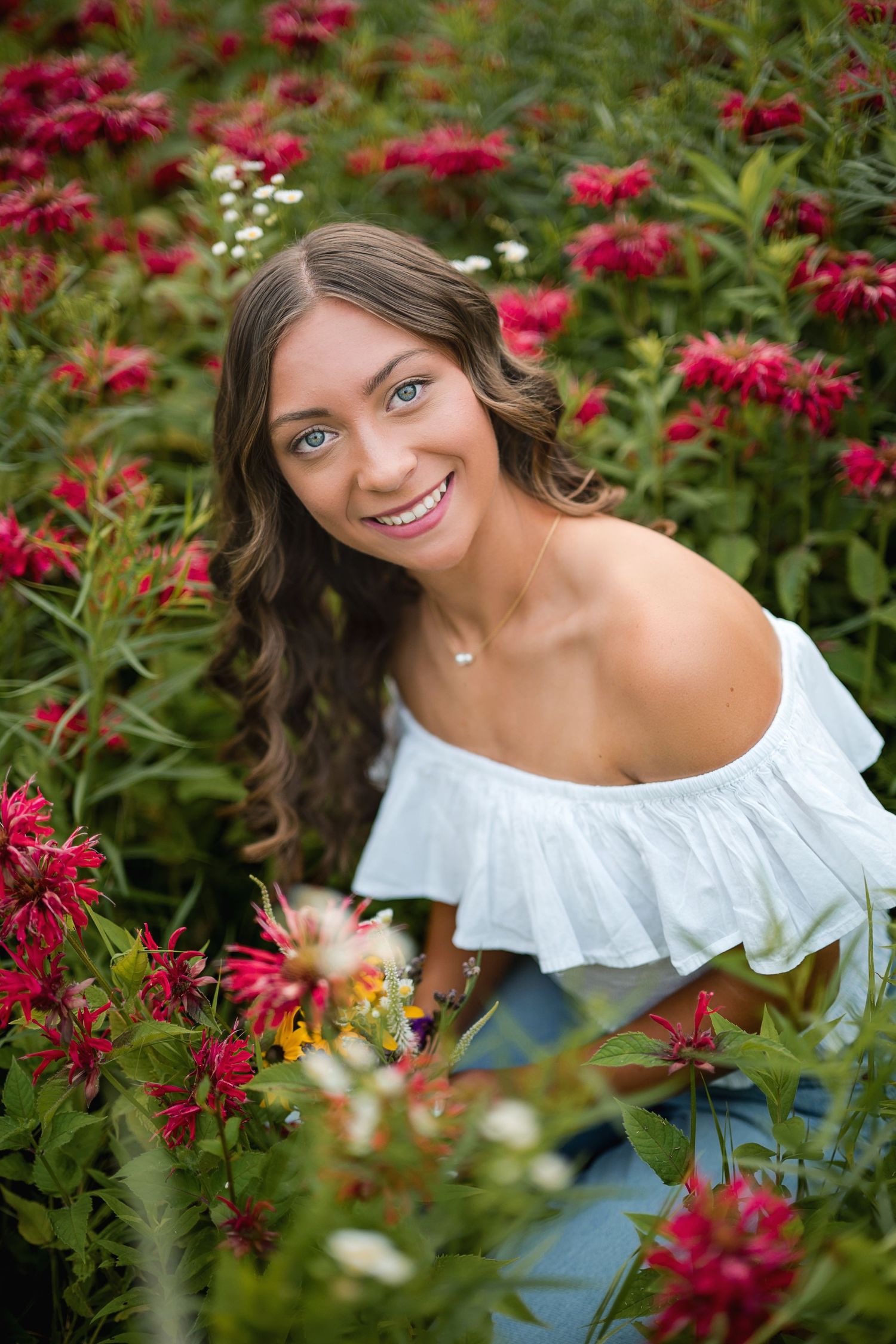 Smiling young woman with long wavy brown hair in a white off-shoulder top poses among red and white wildflowers in a garden.