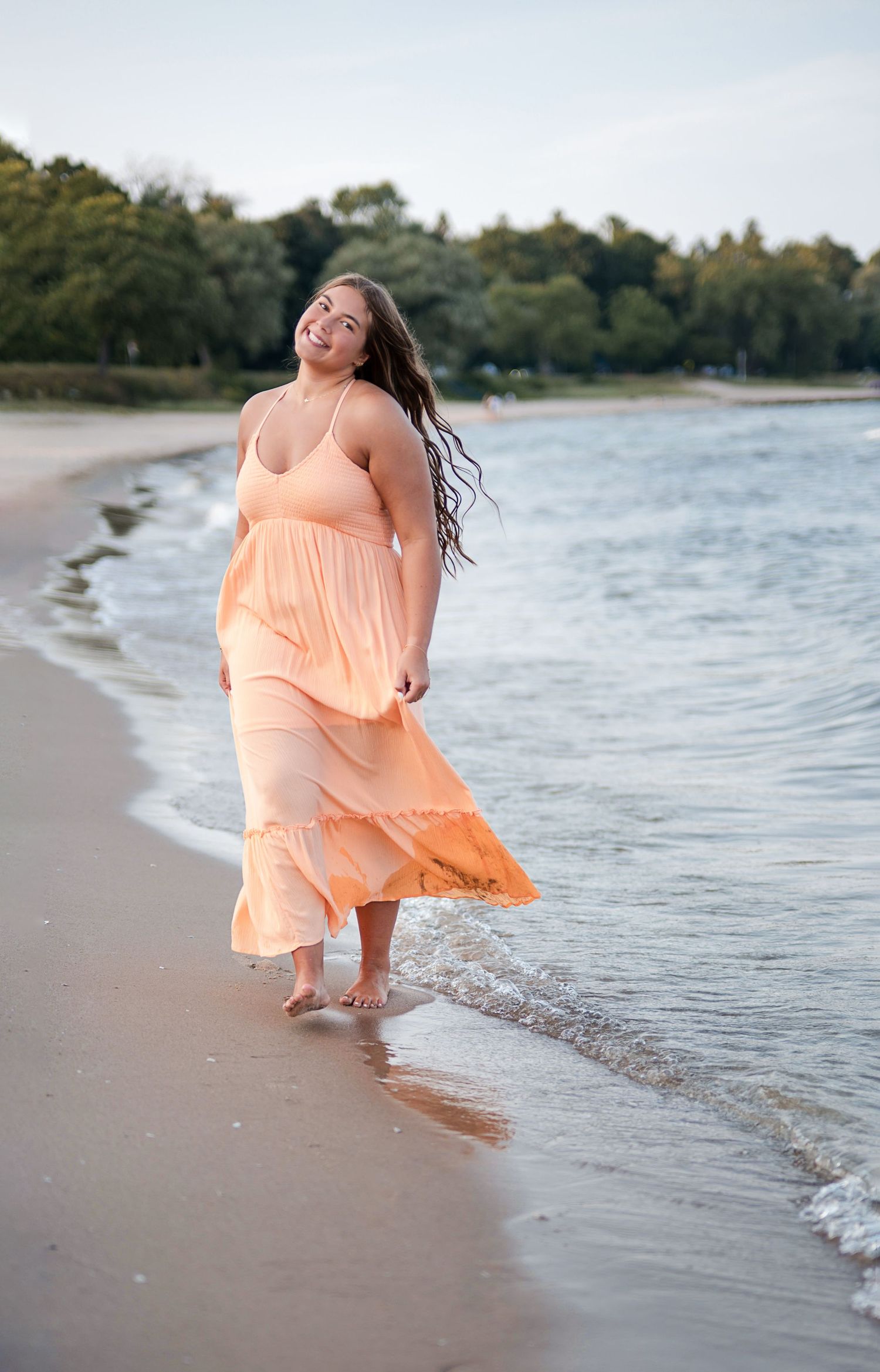 A smiling woman in a flowing peach maxi dress walks barefoot along a sandy beach shoreline with trees in the background.