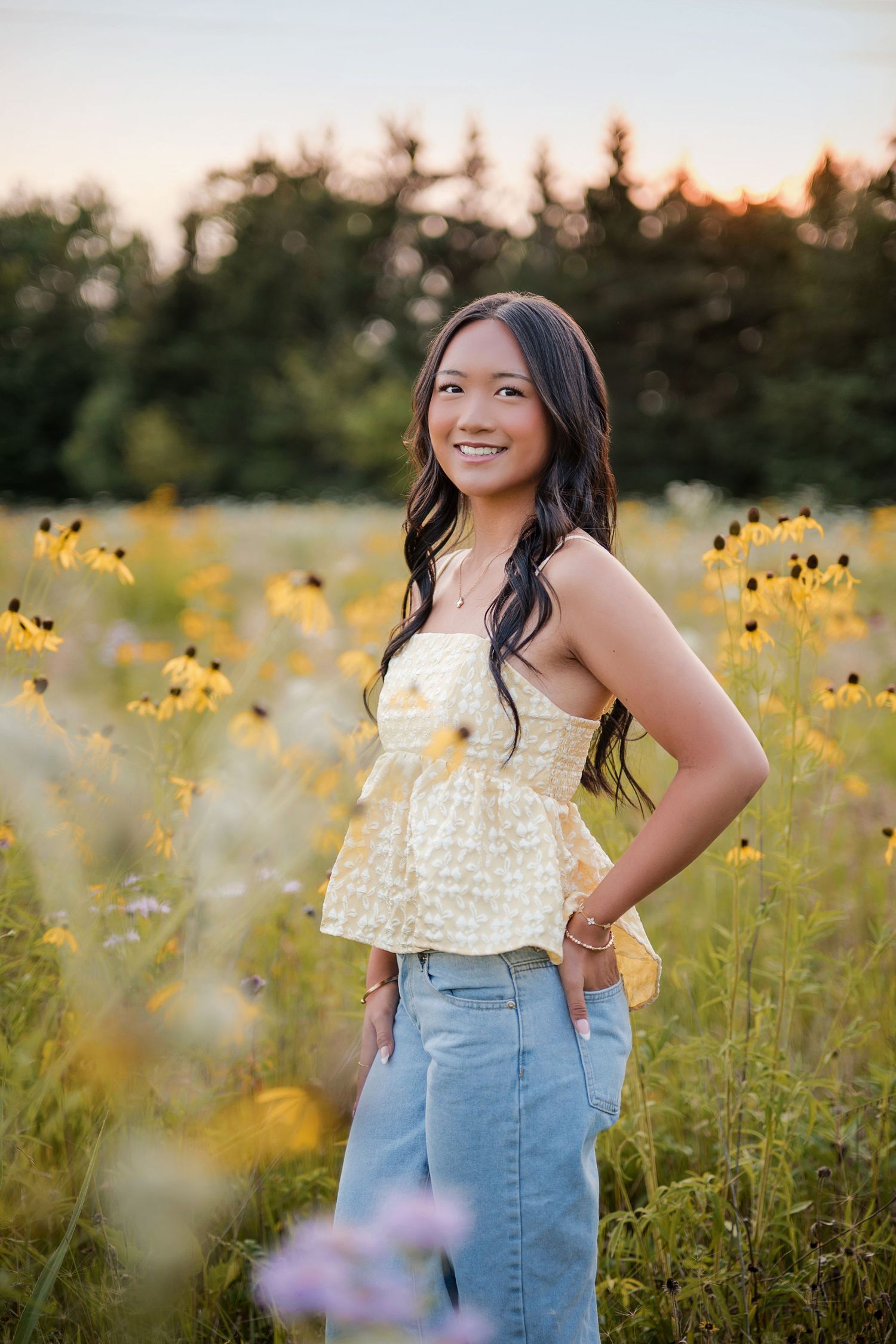 Smiling young woman with long wavy hair in a yellow floral top and jeans stands in a sunset-lit field of yellow wildflowers.
