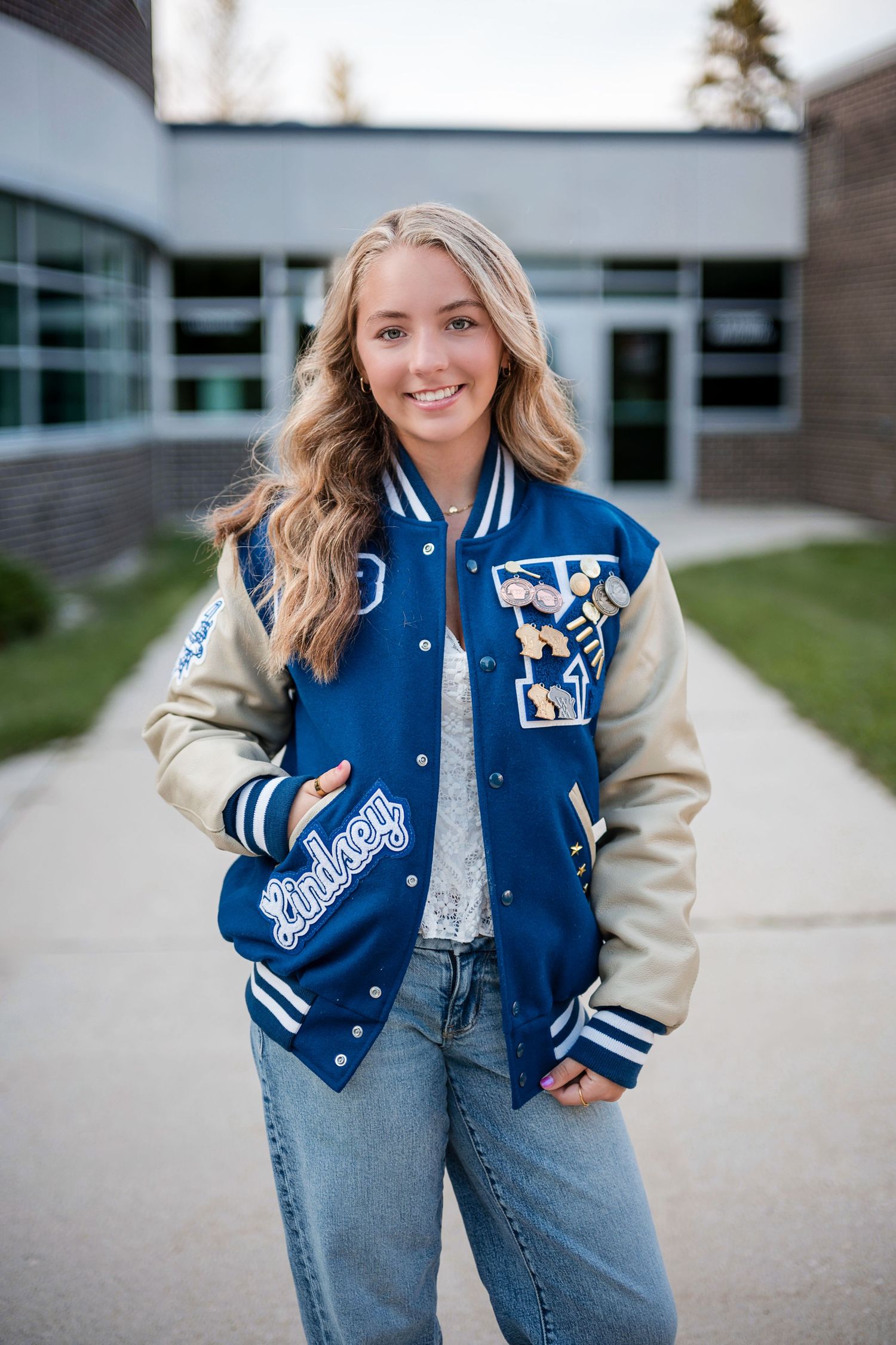 Smiling senior girl in a blue and cream letterman jacket with her name stitched on it, standing outside a school building.