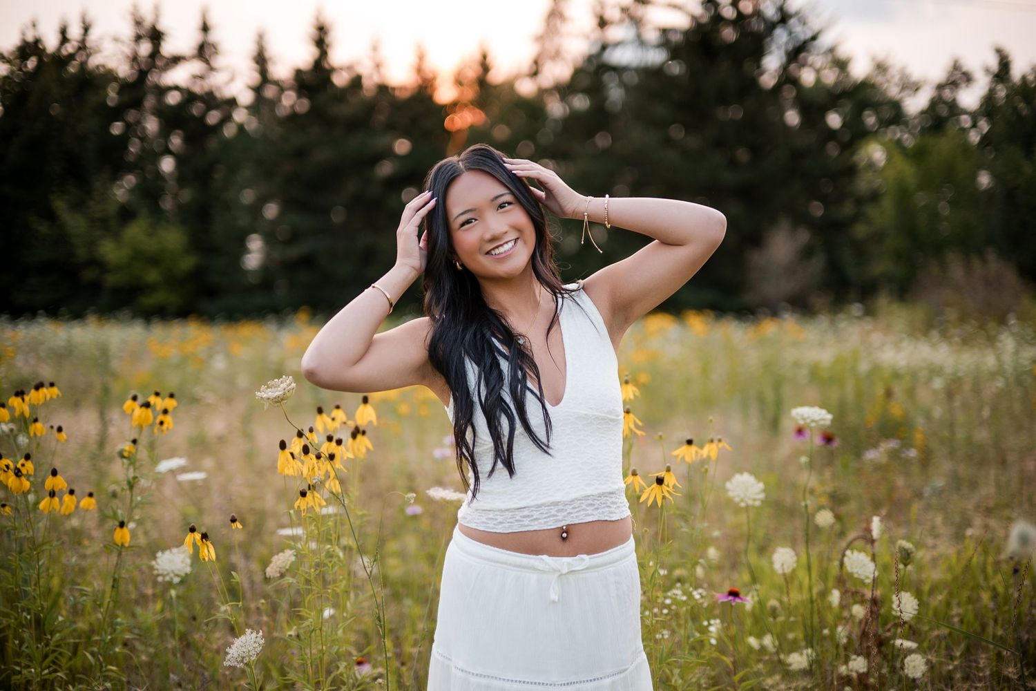 Smiling young woman with long dark hair and a white outfit poses with hands in her hair in a golden wildflower field.