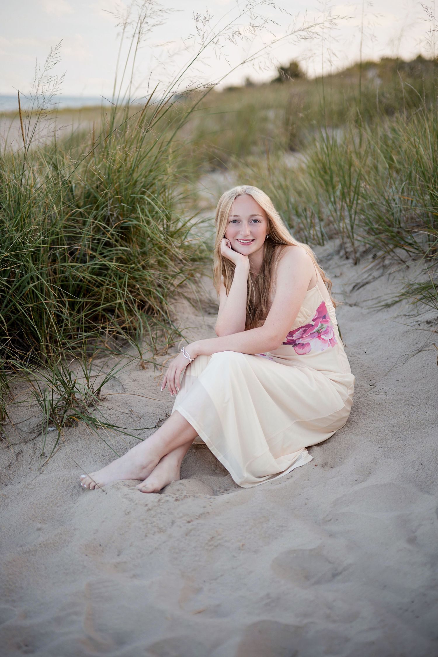 Young blonde woman in floral dress sitting barefoot on sandy beach dunes, smiling with hand on chin.