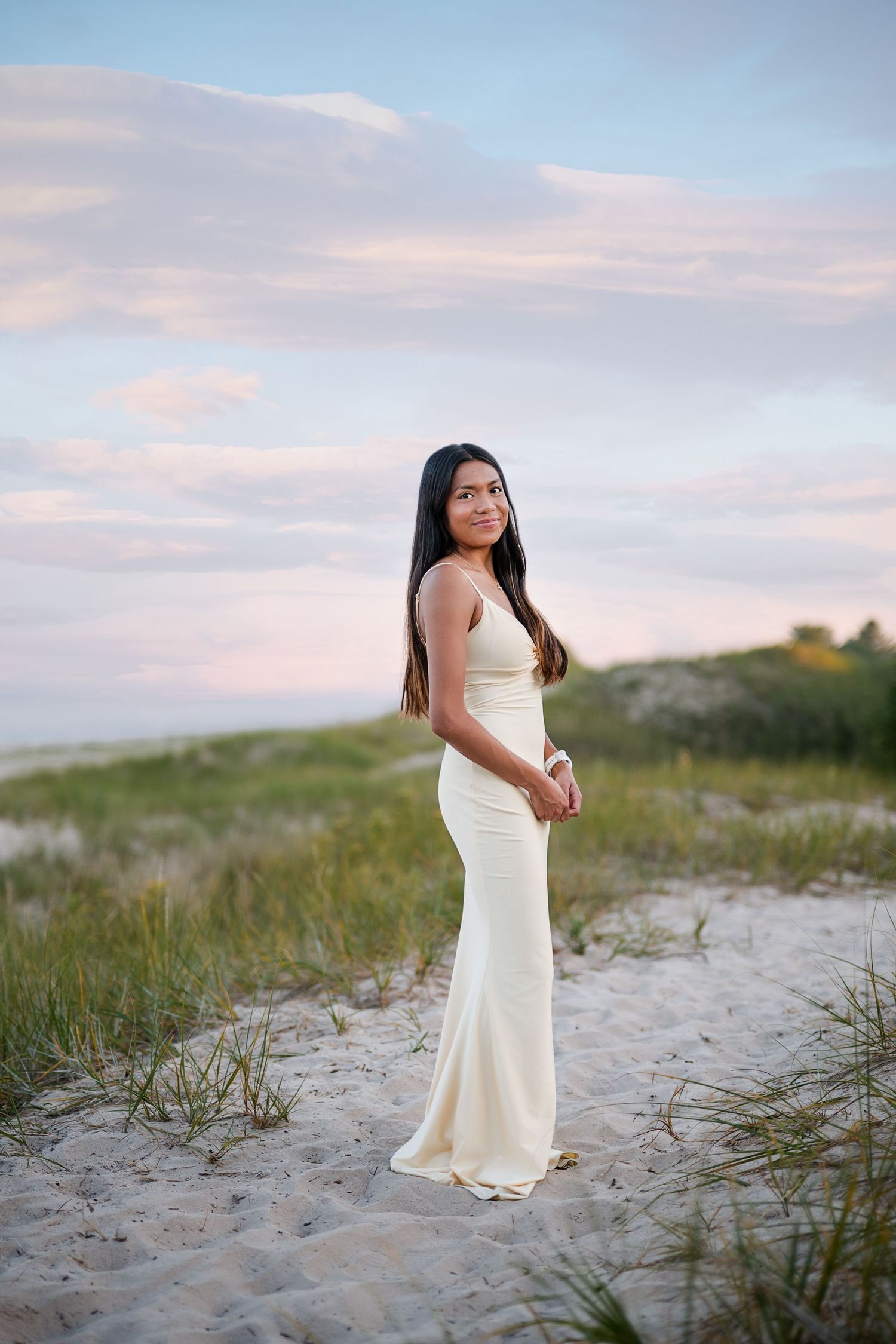 Young woman in elegant cream gown standing on sandy beach dunes at sunset with soft pastel sky.