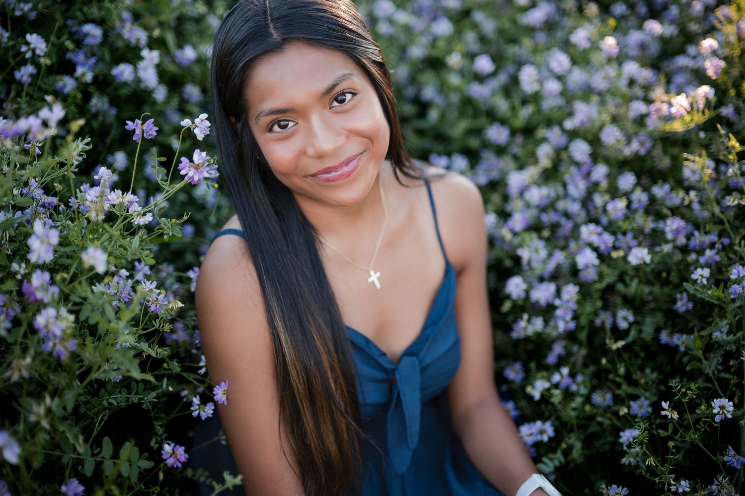 Young woman with long dark hair wearing a teal dress and cross necklace, smiling among purple flowering bushes.