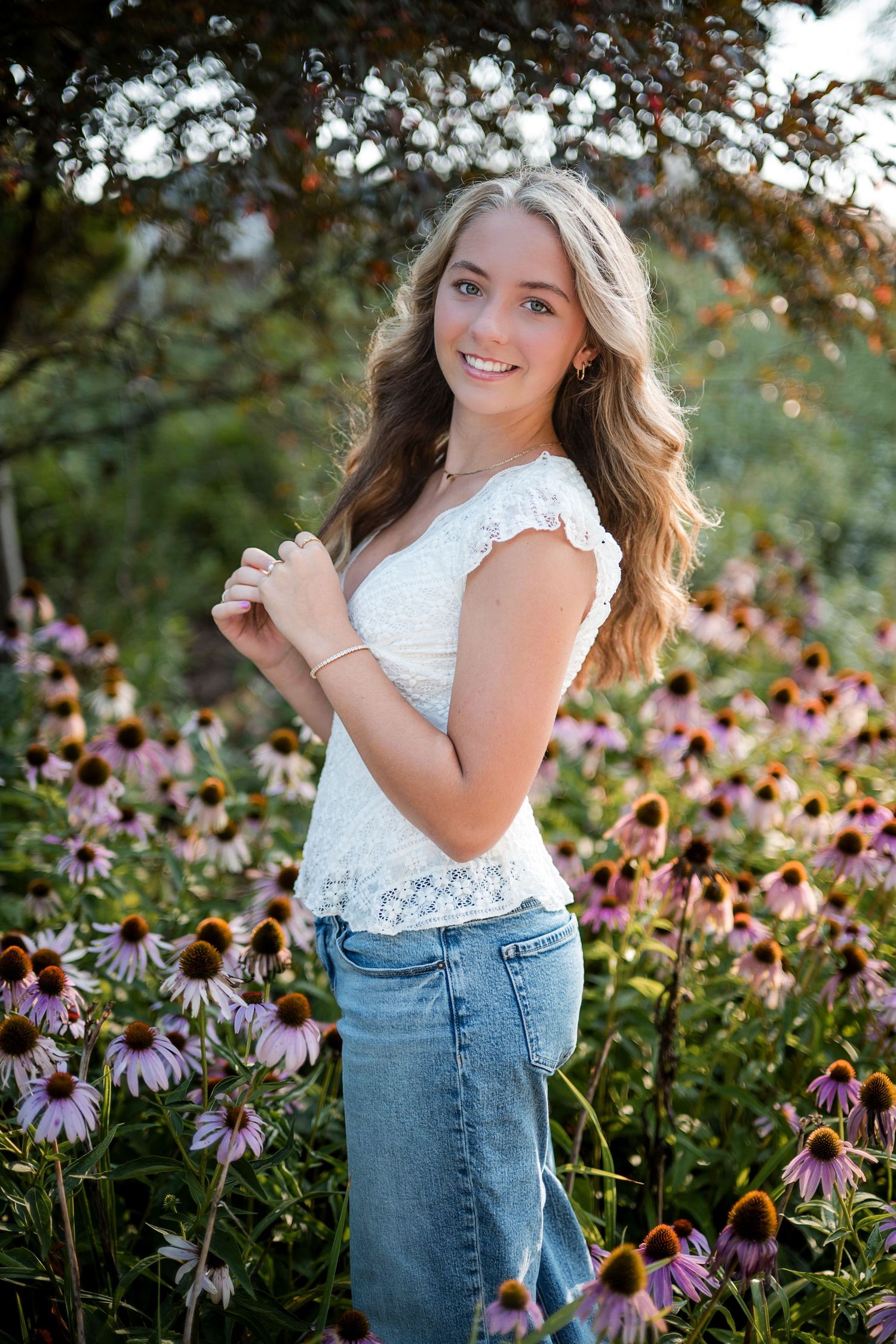 Smiling young woman with long wavy hair in a white lace top and jeans standing among purple coneflowers in a garden.