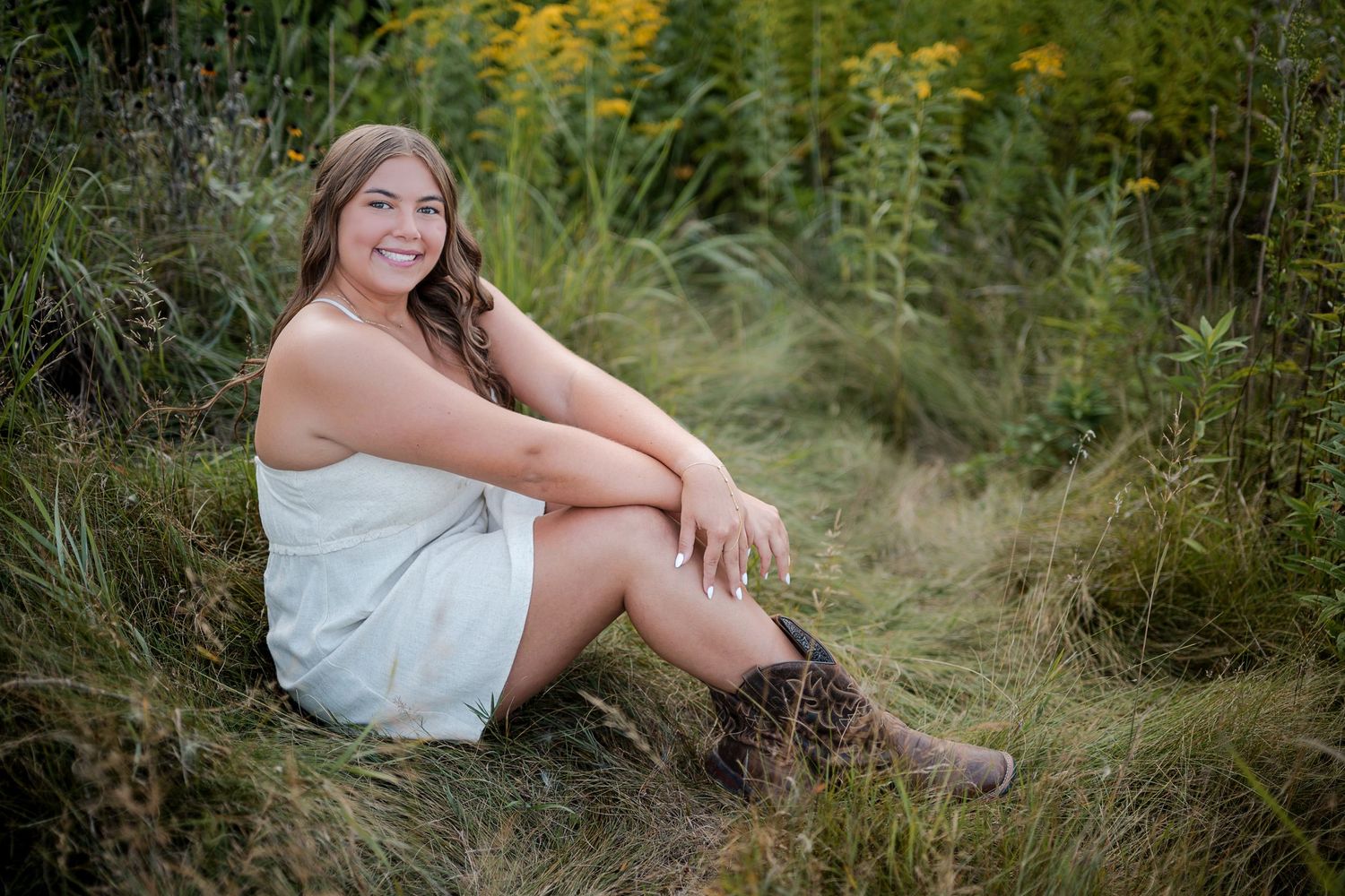 Young woman in white dress and cowboy boots sitting in a meadow with wildflowers and tall grass.