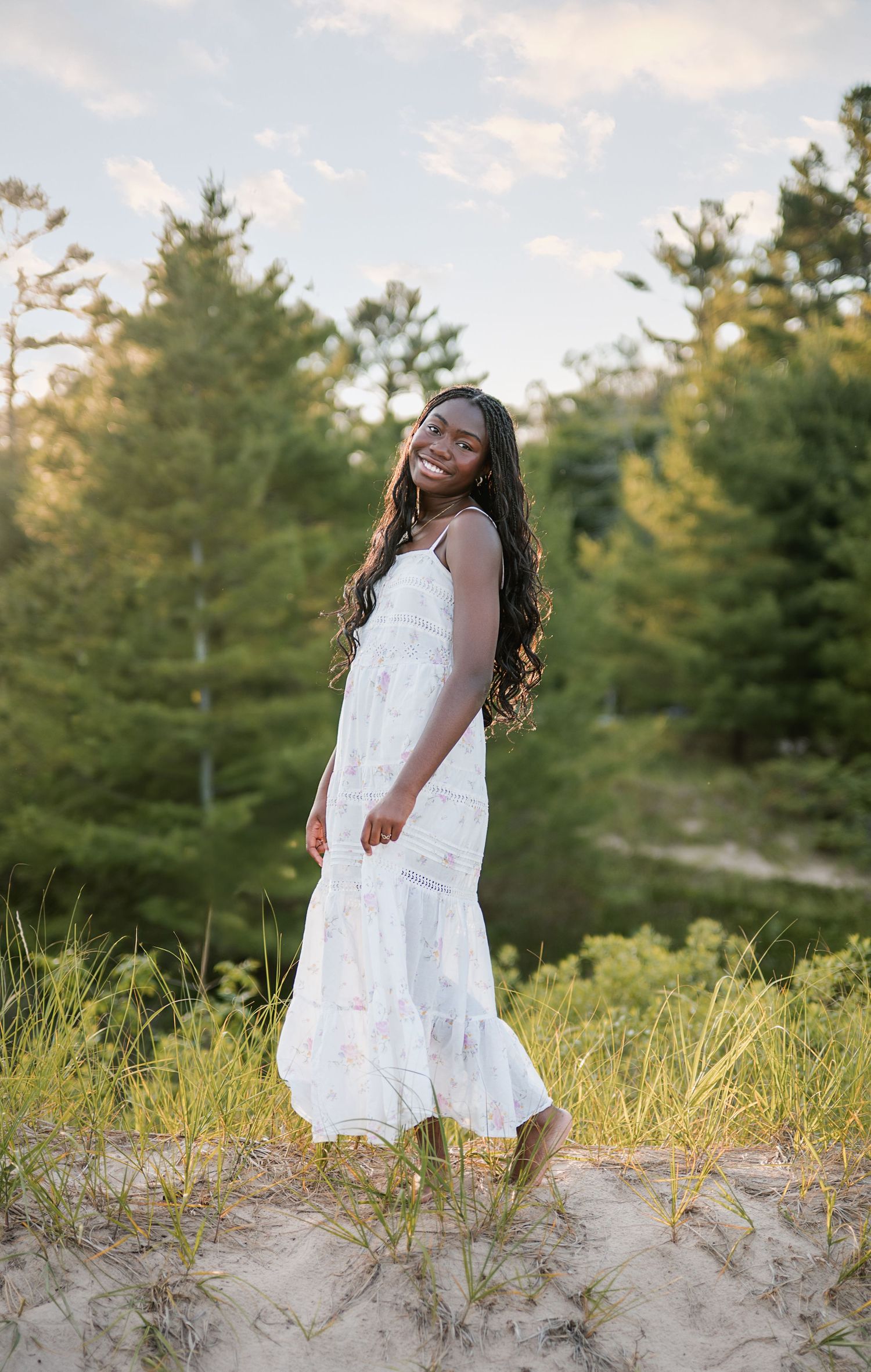 Smiling young woman in a white sundress with long curly hair poses on a sandy dune in front of green pine trees.