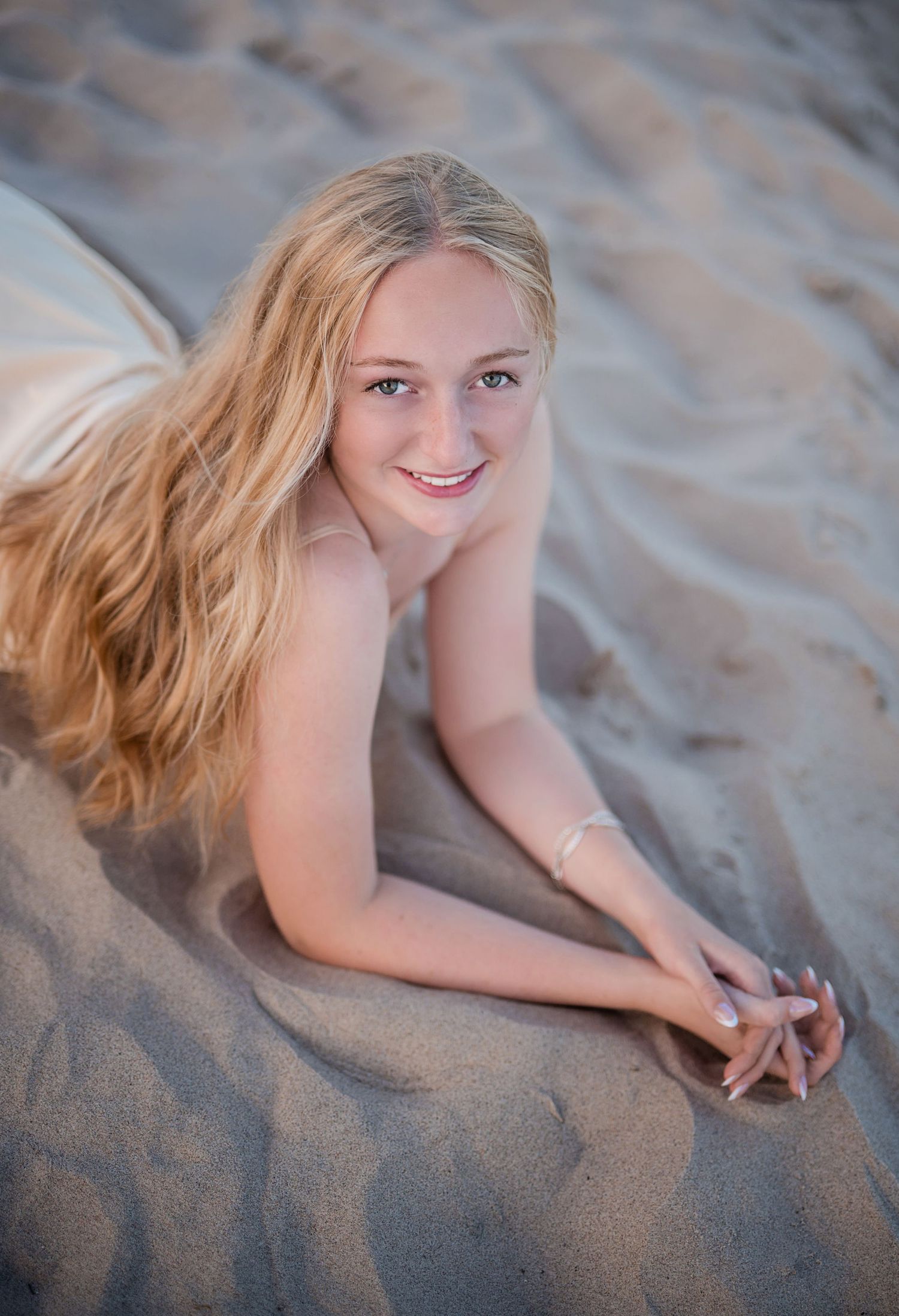 Young blonde woman smiling while lying on sandy beach, looking up at camera on a sunny day.