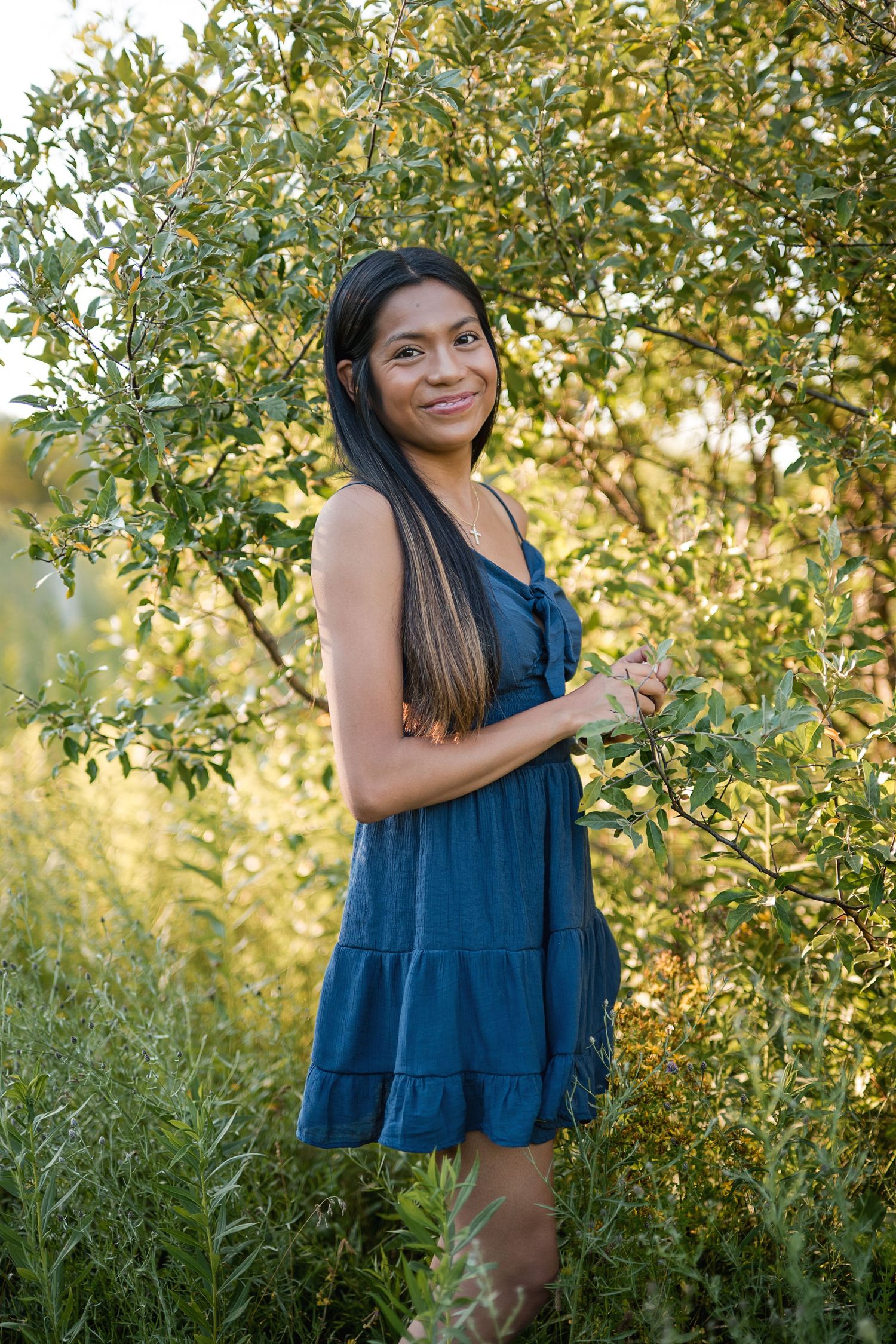 Smiling young woman in blue sundress stands among lush green trees outdoors.