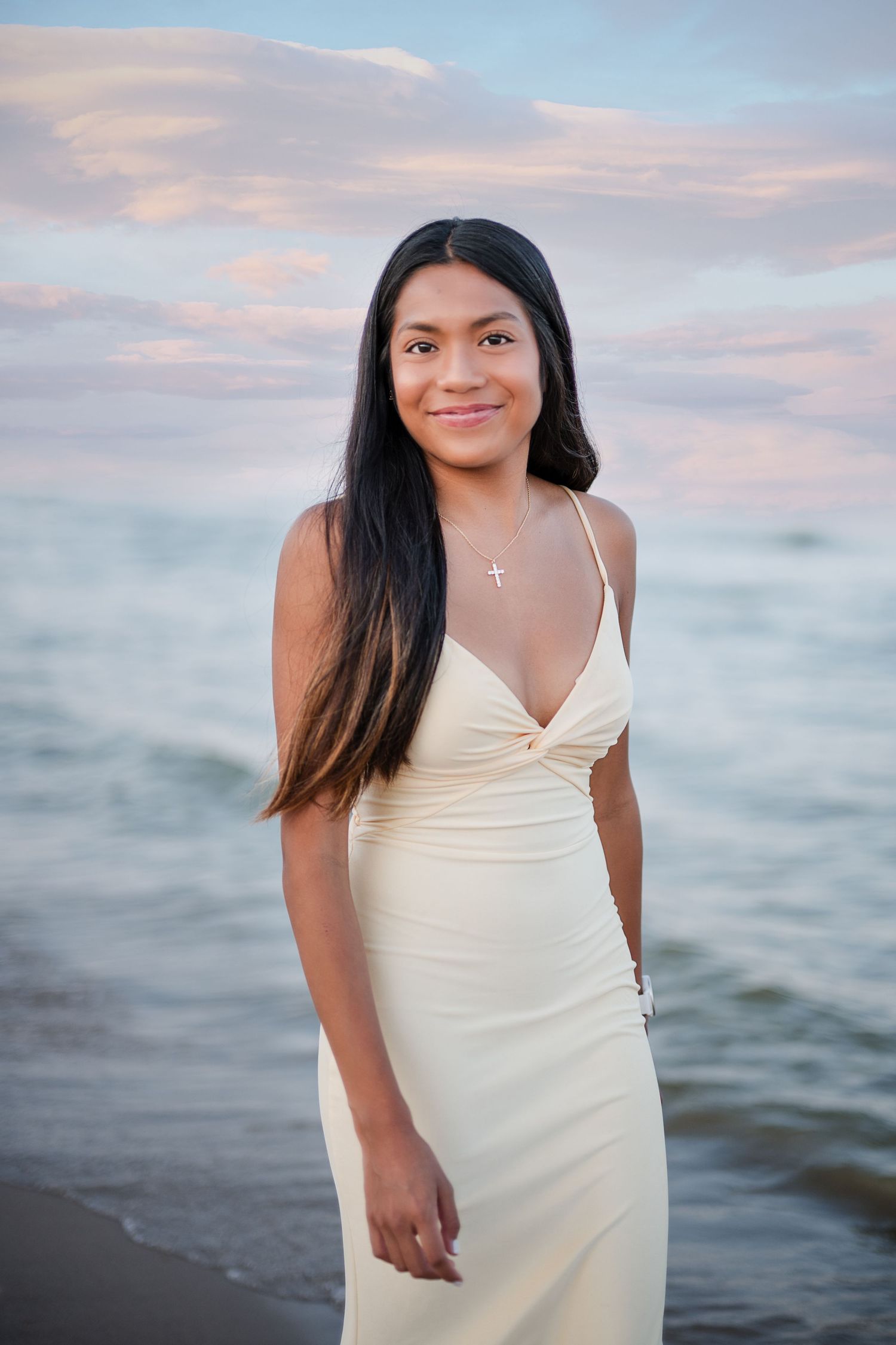 Young woman in cream dress with cross necklace smiling at beach during sunset.