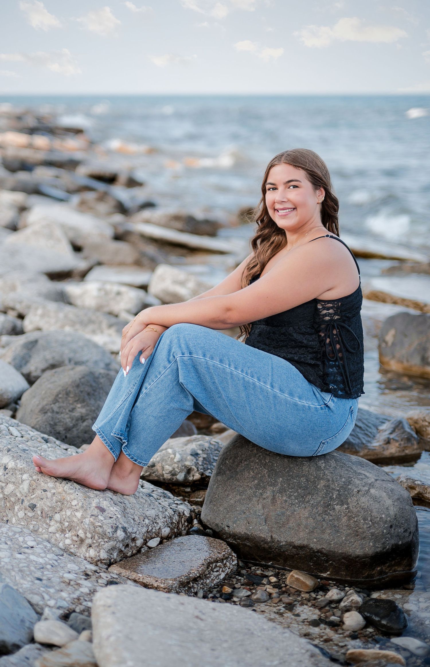 Young woman in jeans and black top sitting barefoot on rocks by the ocean, smiling at camera.