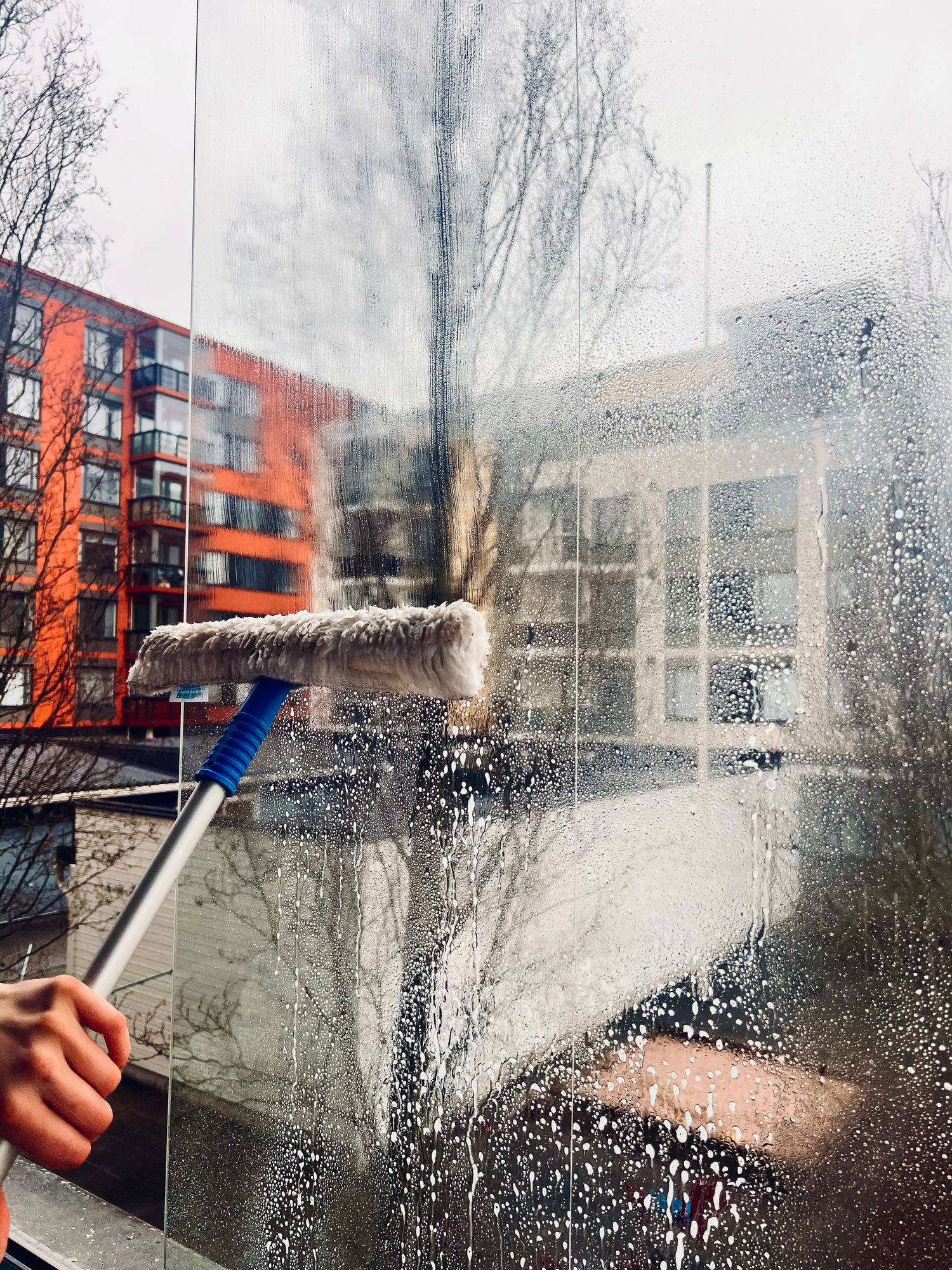 Person cleaning large glass window with squeegee mop on urban street with colorful buildings reflected.