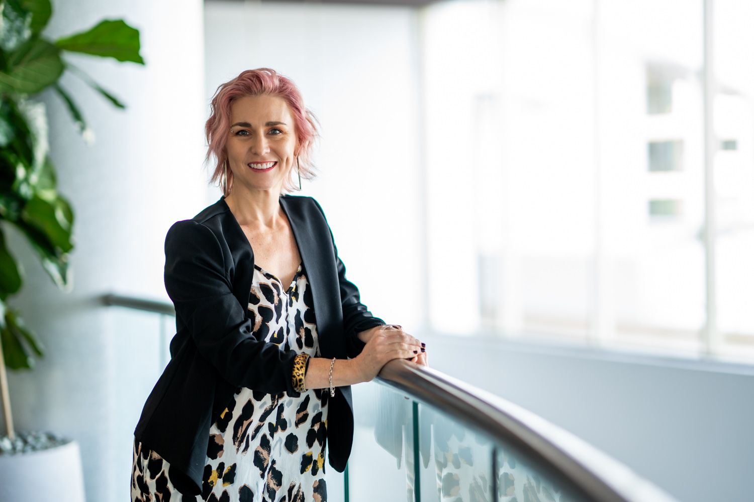 A woman with pink hair in a black blazer and animal print dress smiles while leaning on a railing indoors.