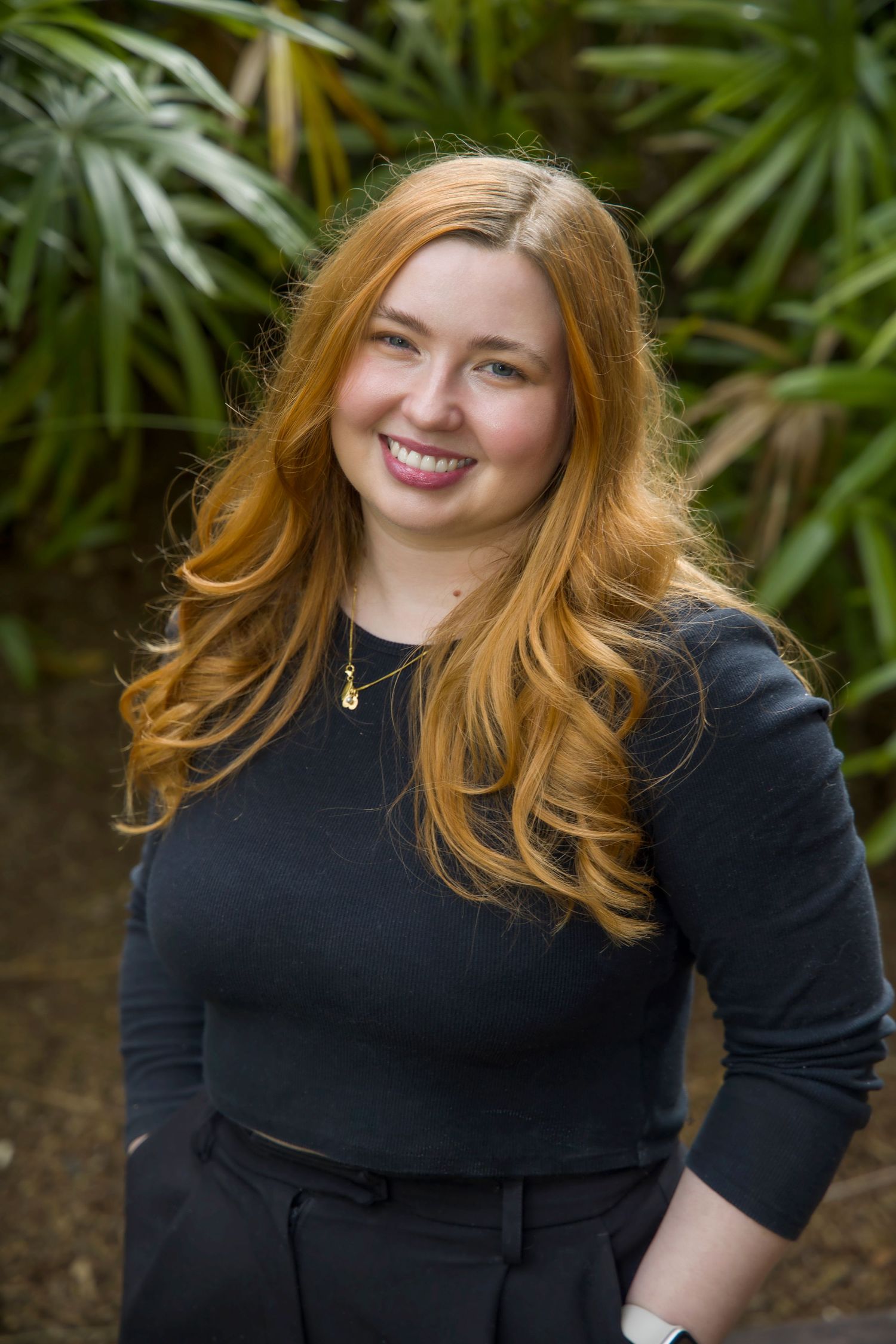 Young woman with long auburn hair in black top smiling outdoors surrounded by tropical green foliage.