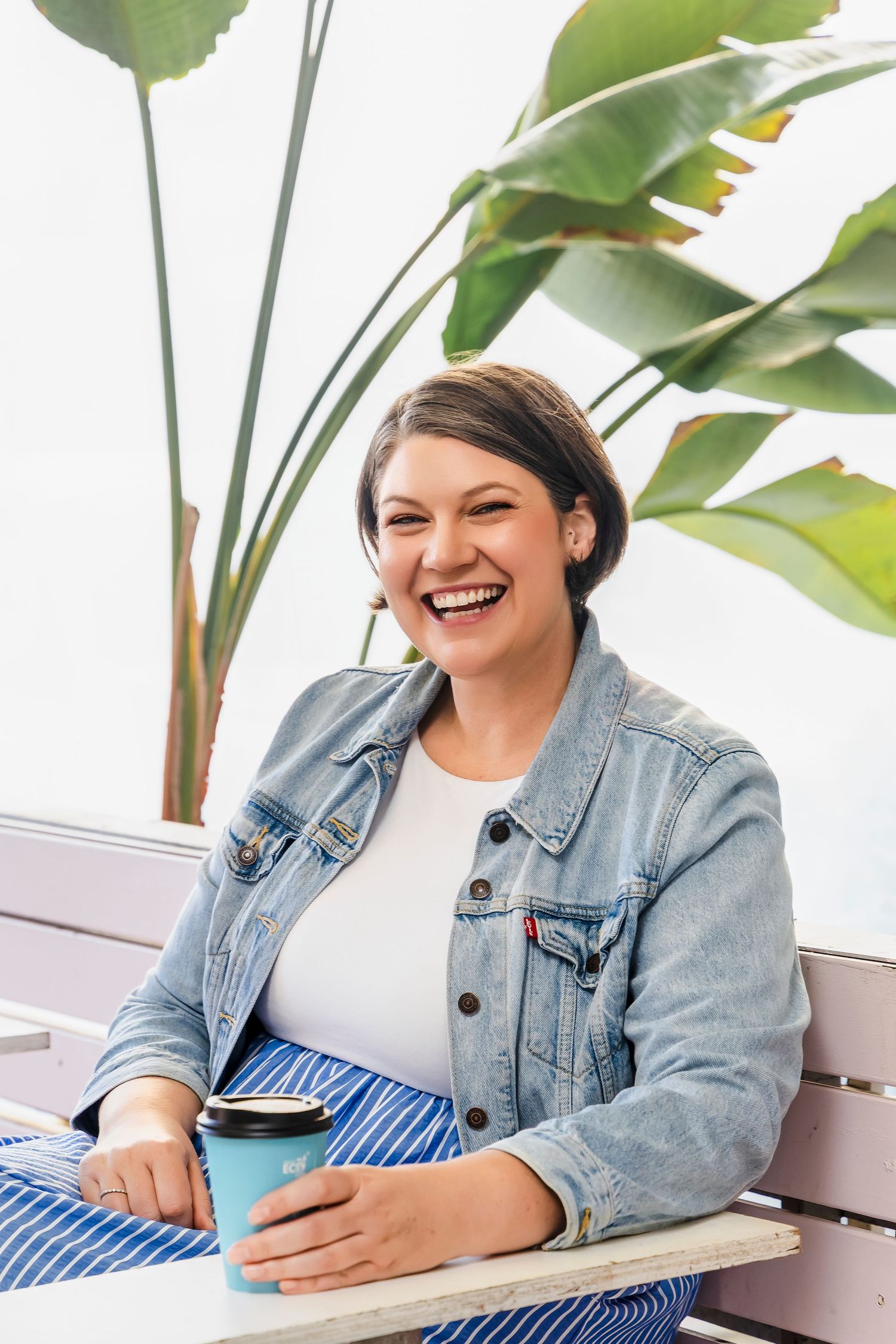 Smiling woman in denim jacket holding a coffee cup seated near a tropical plant.