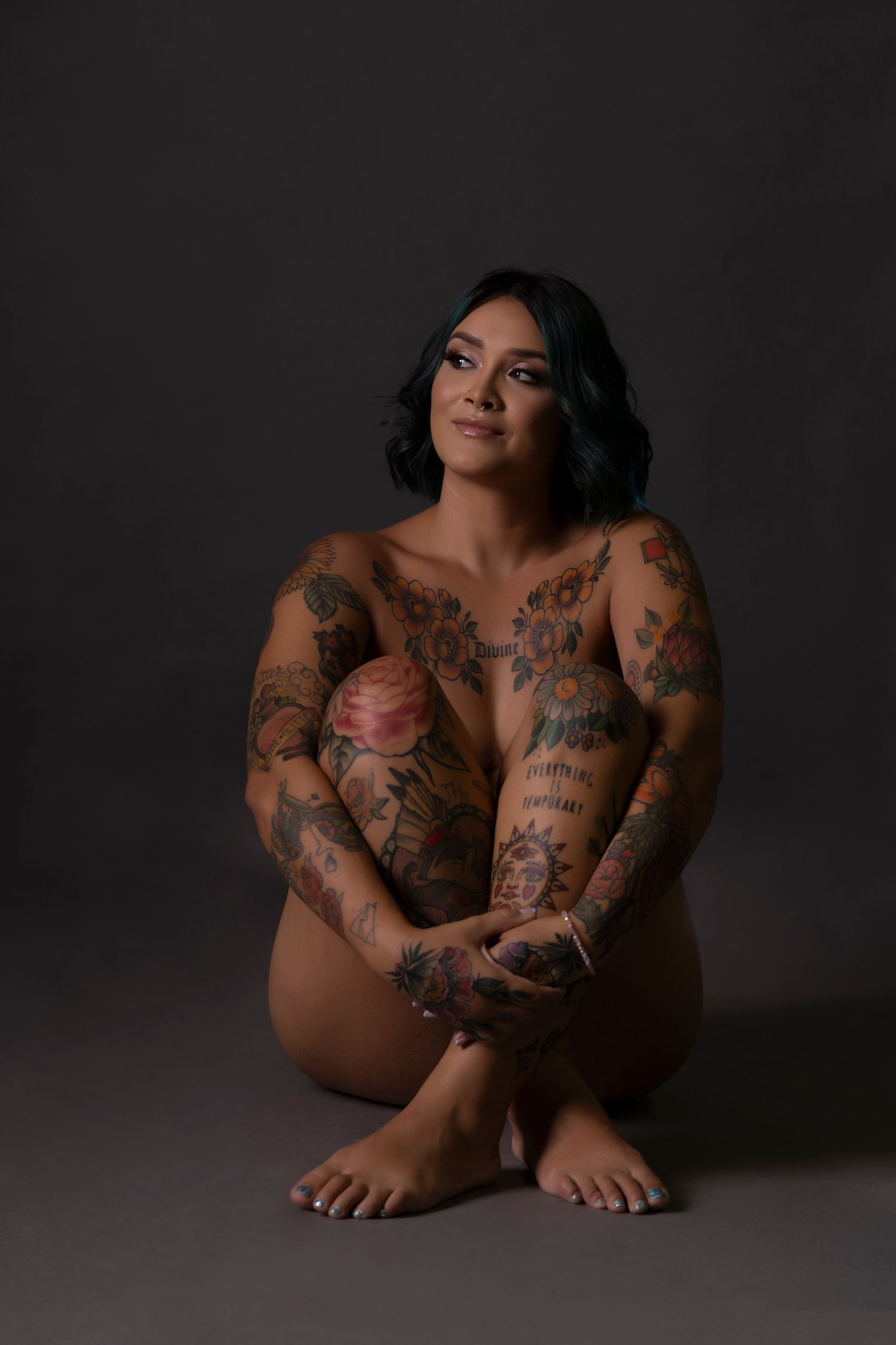 Heavily tattooed woman sitting cross-legged on floor against dark gray studio background.