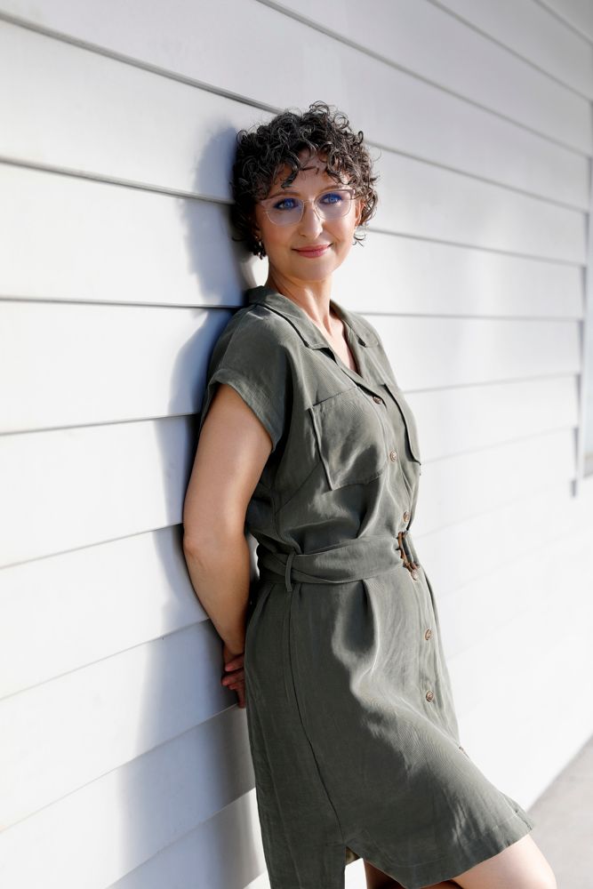 A smiling curly-haired woman in a green dress leans against a white paneled wall.