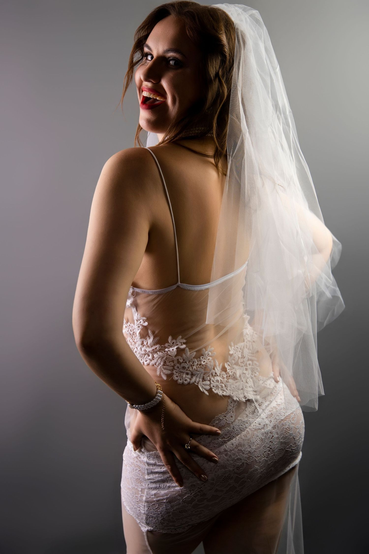 Woman in white lace bridal lingerie and veil posing with back to camera on gray background.