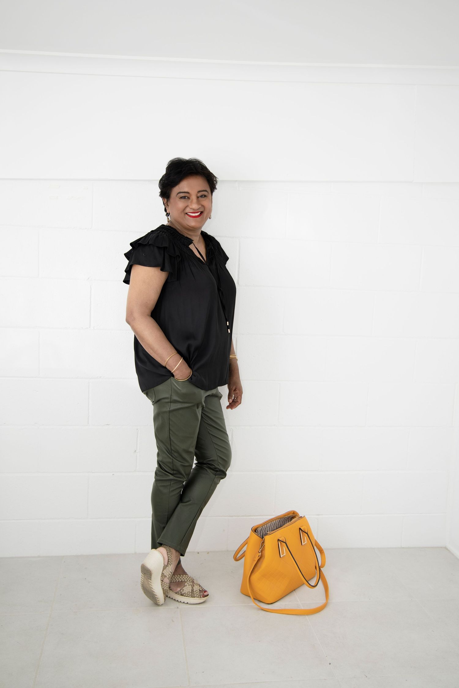 Stylish woman in black blouse and olive pants leaning against white wall with yellow leather handbag.