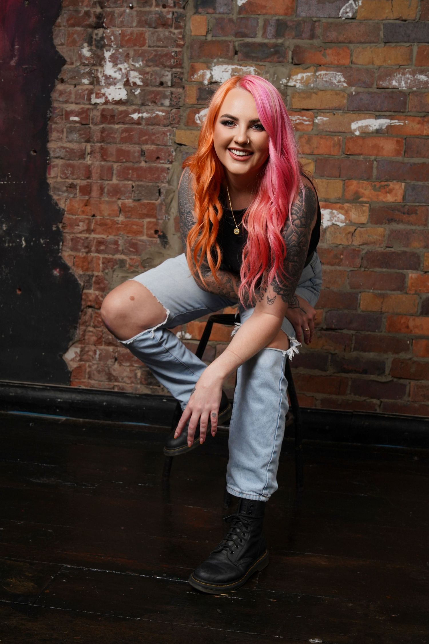 Woman with pink and orange hair in ripped jeans and boots poses against brick wall.
