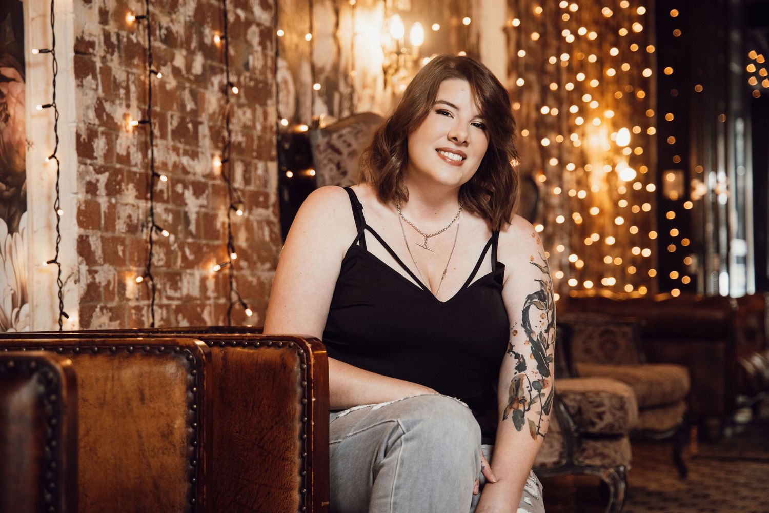 Tattooed woman in black tank top smiles warmly surrounded by warm bokeh fairy lights.