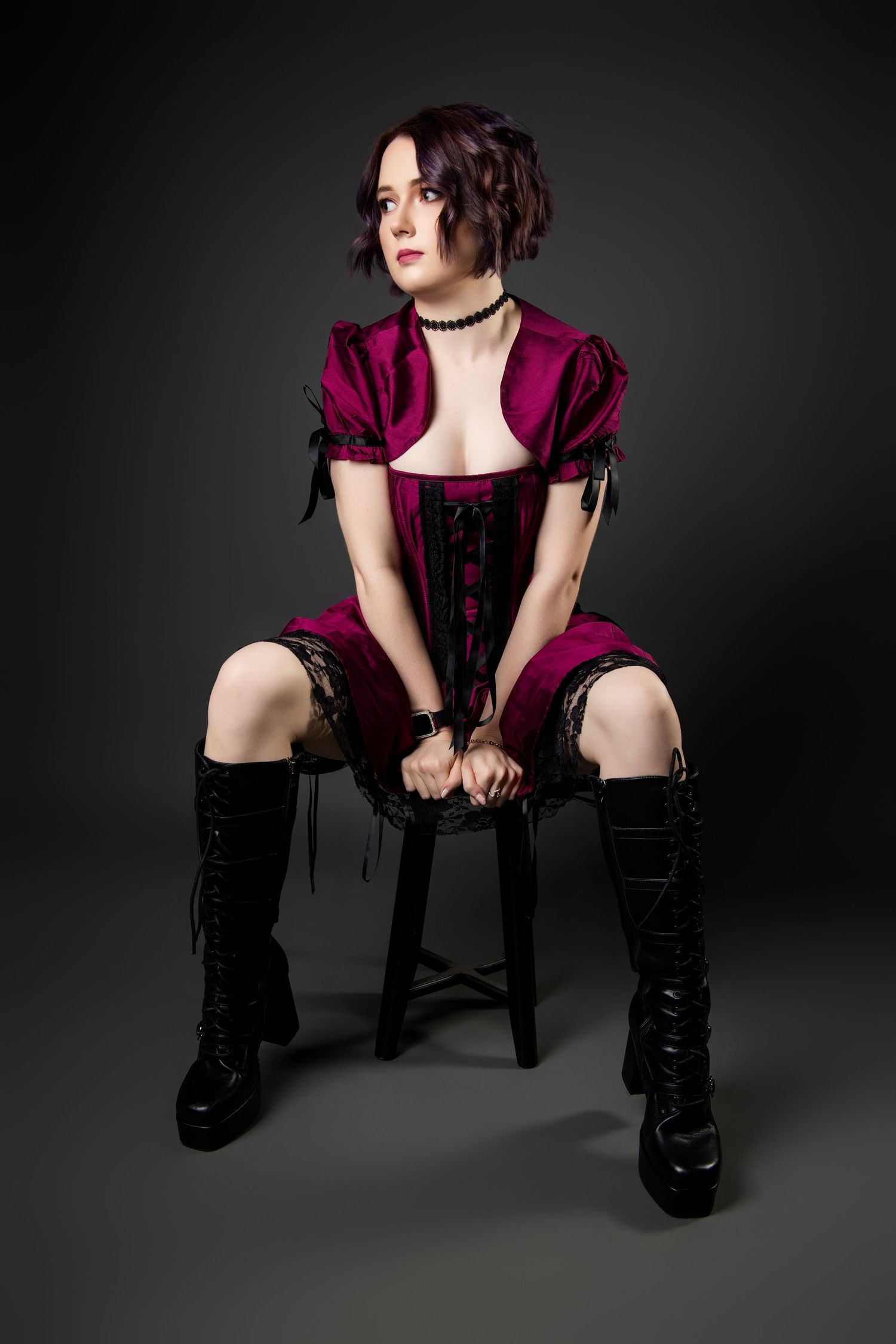 Woman in purple corset dress and black knee-high boots sitting on stool against dark background.