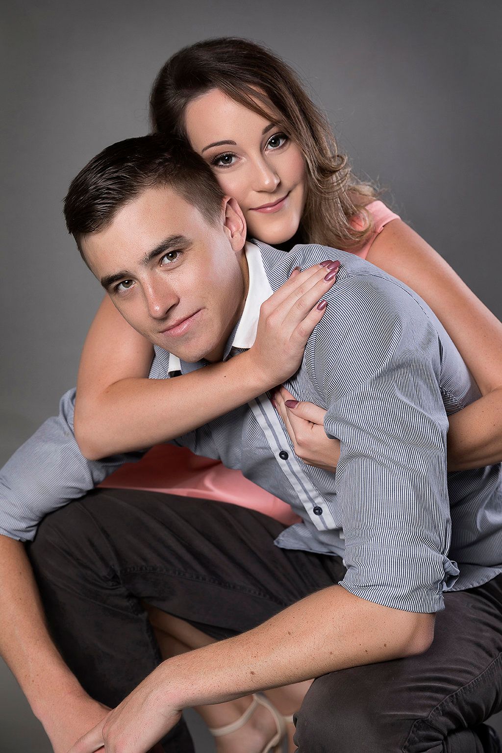 A young couple poses together in a studio, the woman embracing the man from behind affectionately.