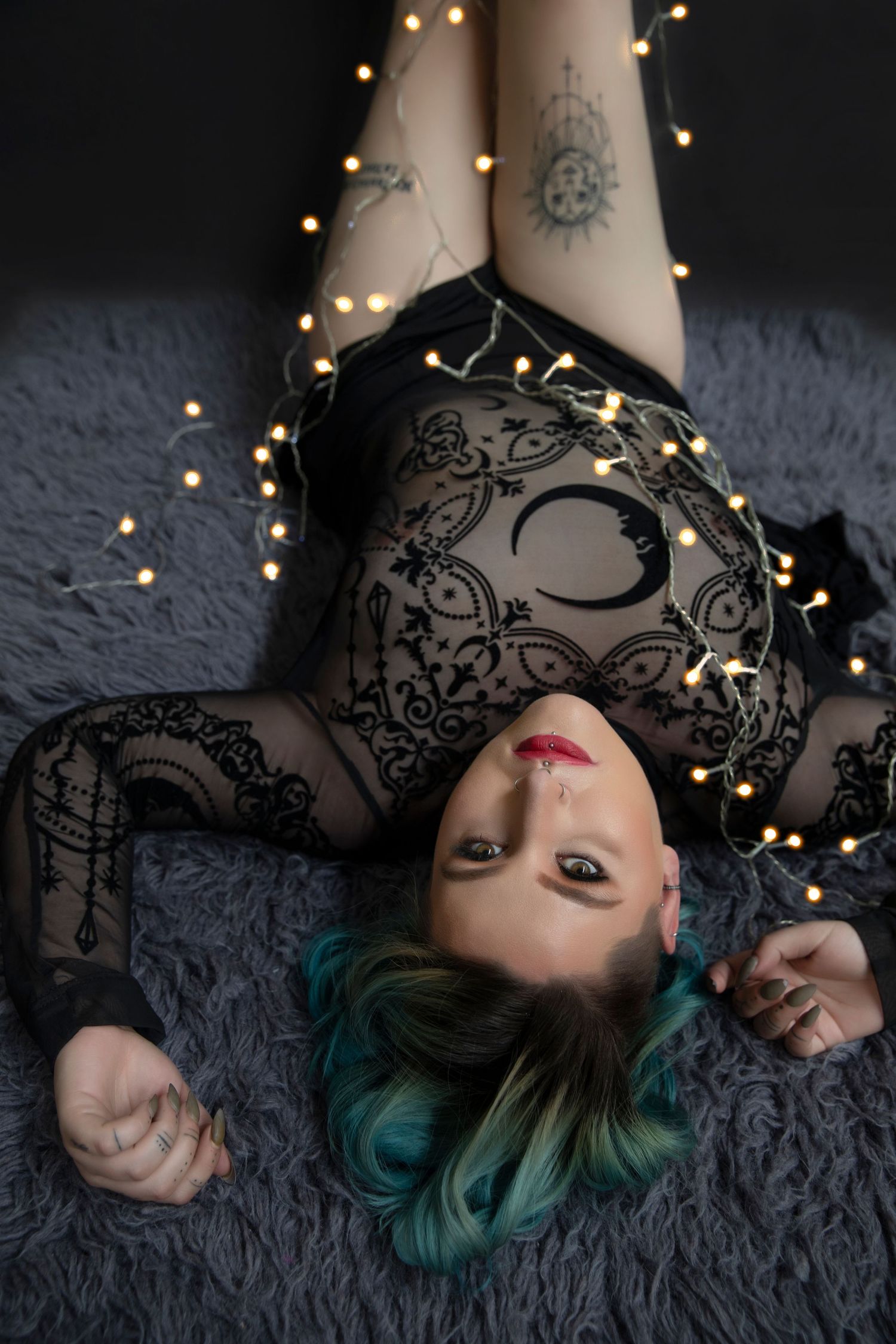 Woman with teal hair lying on fur rug wearing black lace bodysuit surrounded by string lights.