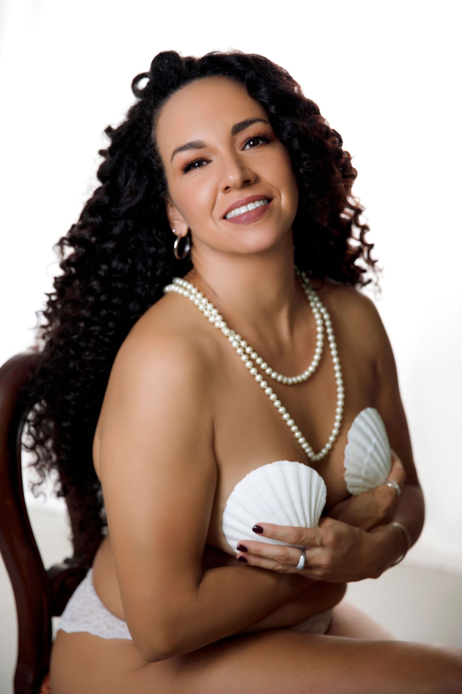 Woman with curly hair smiling in white lingerie holding seashell wearing pearl necklace in boudoir.