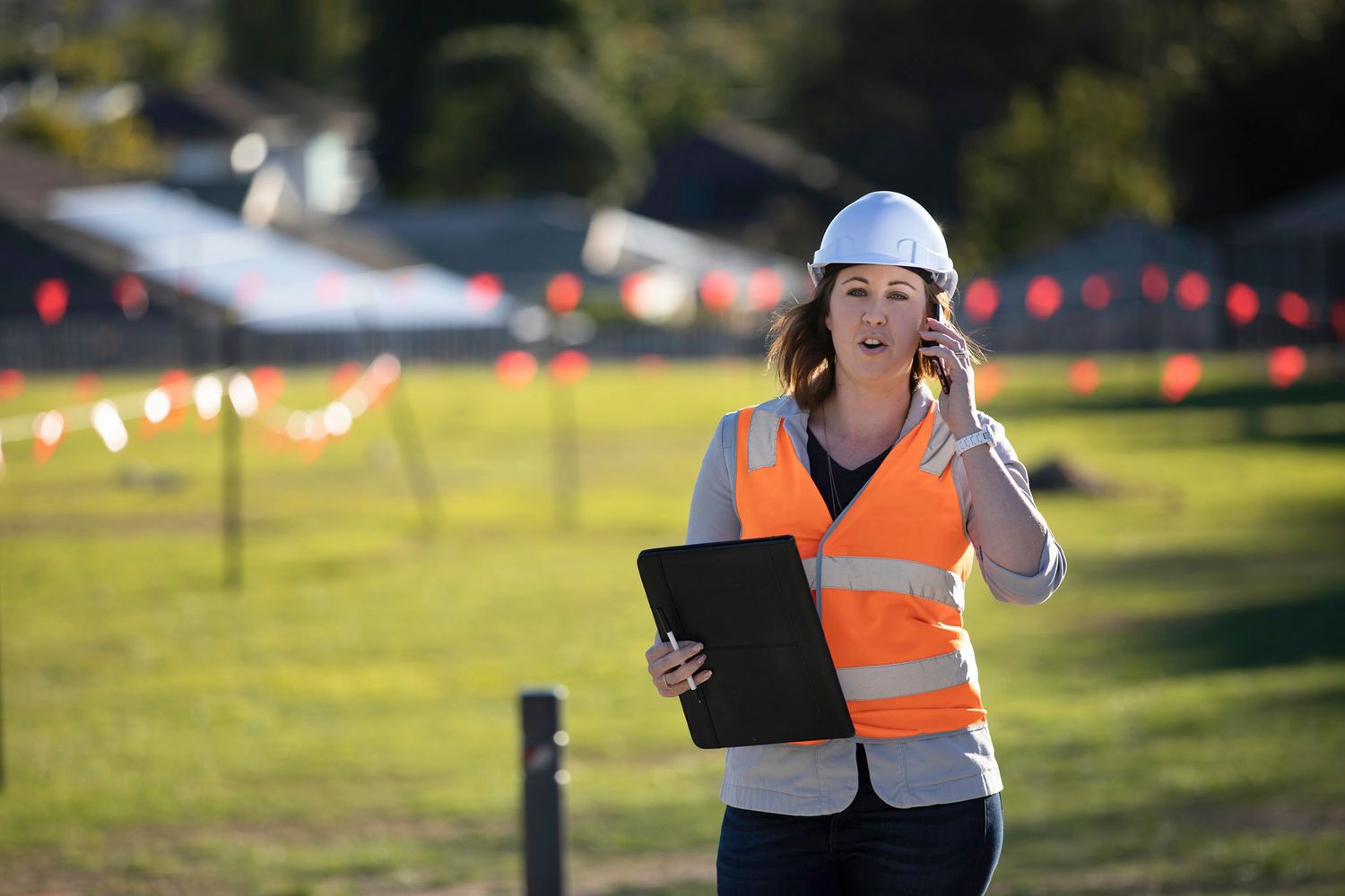 Female engineer in orange vest and hard hat holding clipboard while on phone outdoors.