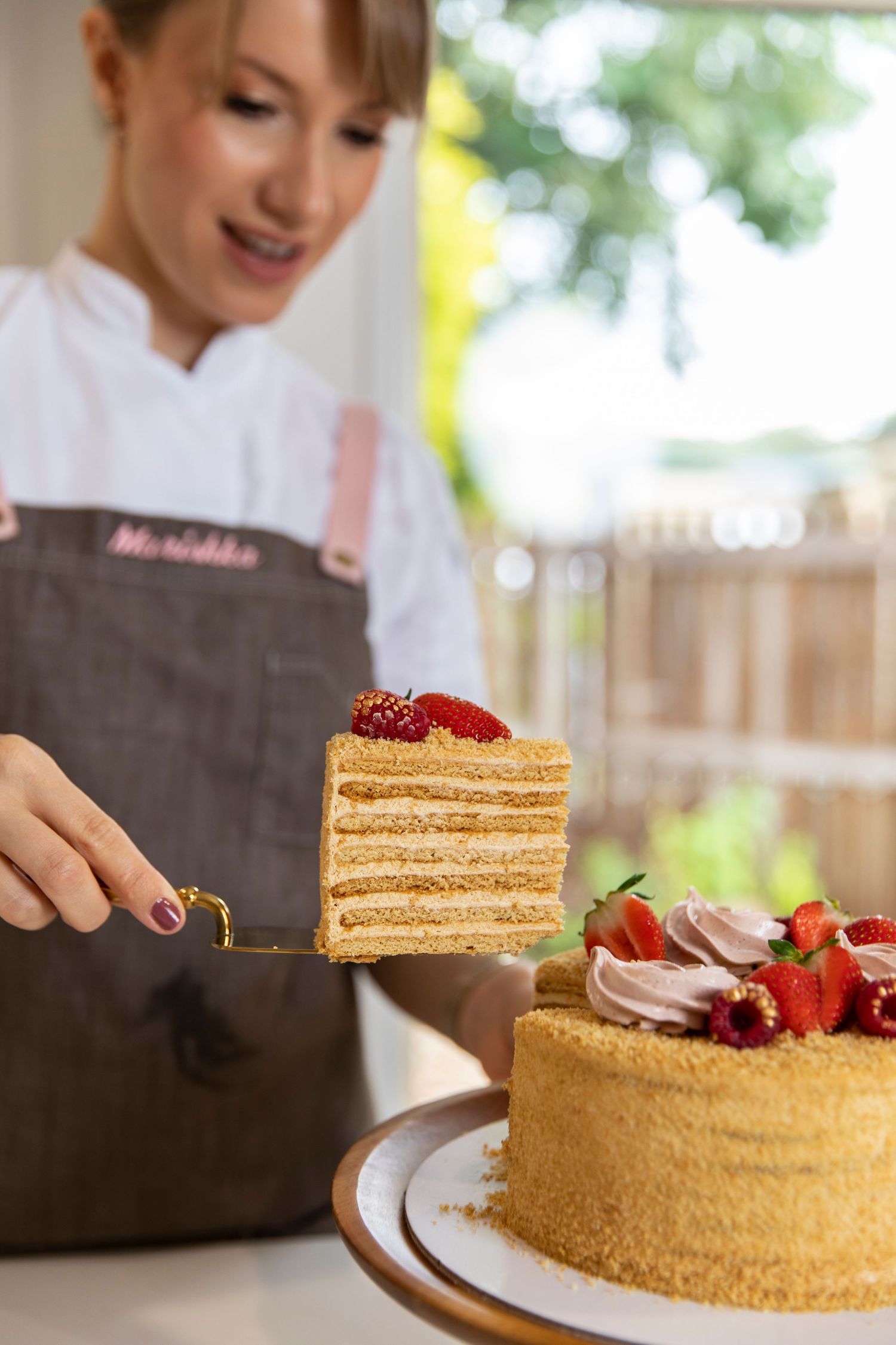 Smiling pastry chef in grey apron serving slice of layered honey cake with strawberries.