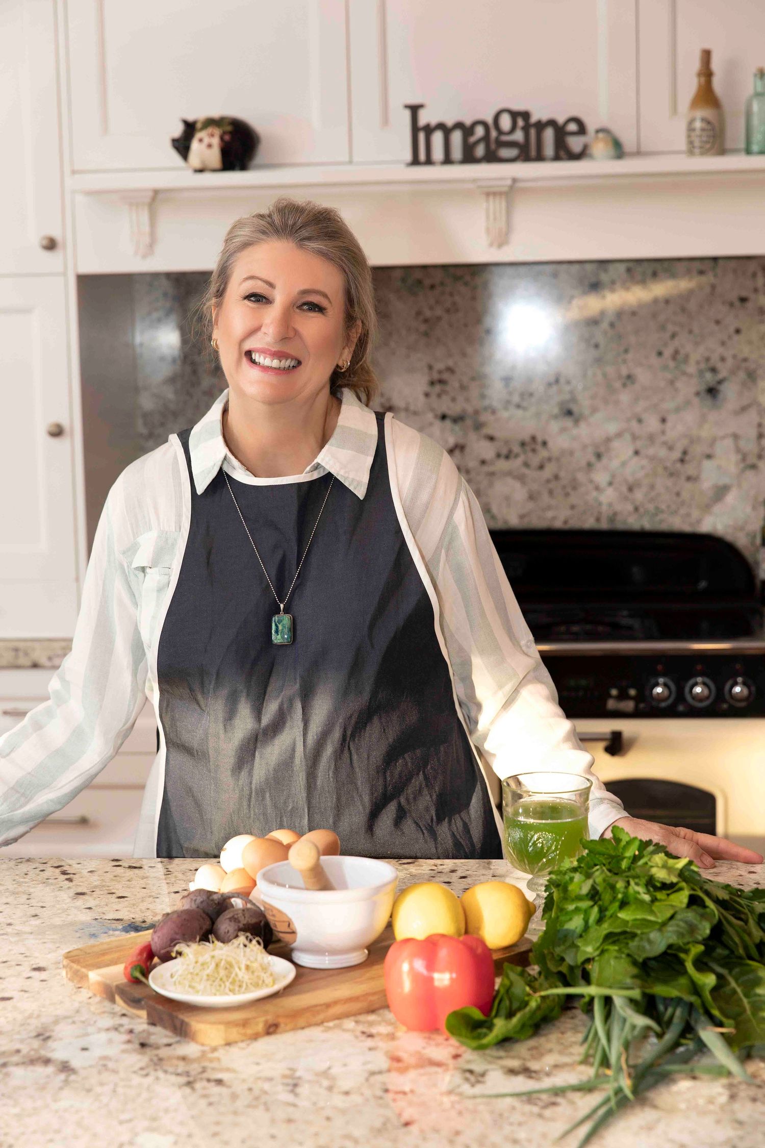 Smiling woman in apron standing at kitchen counter with fresh vegetables and ingredients.